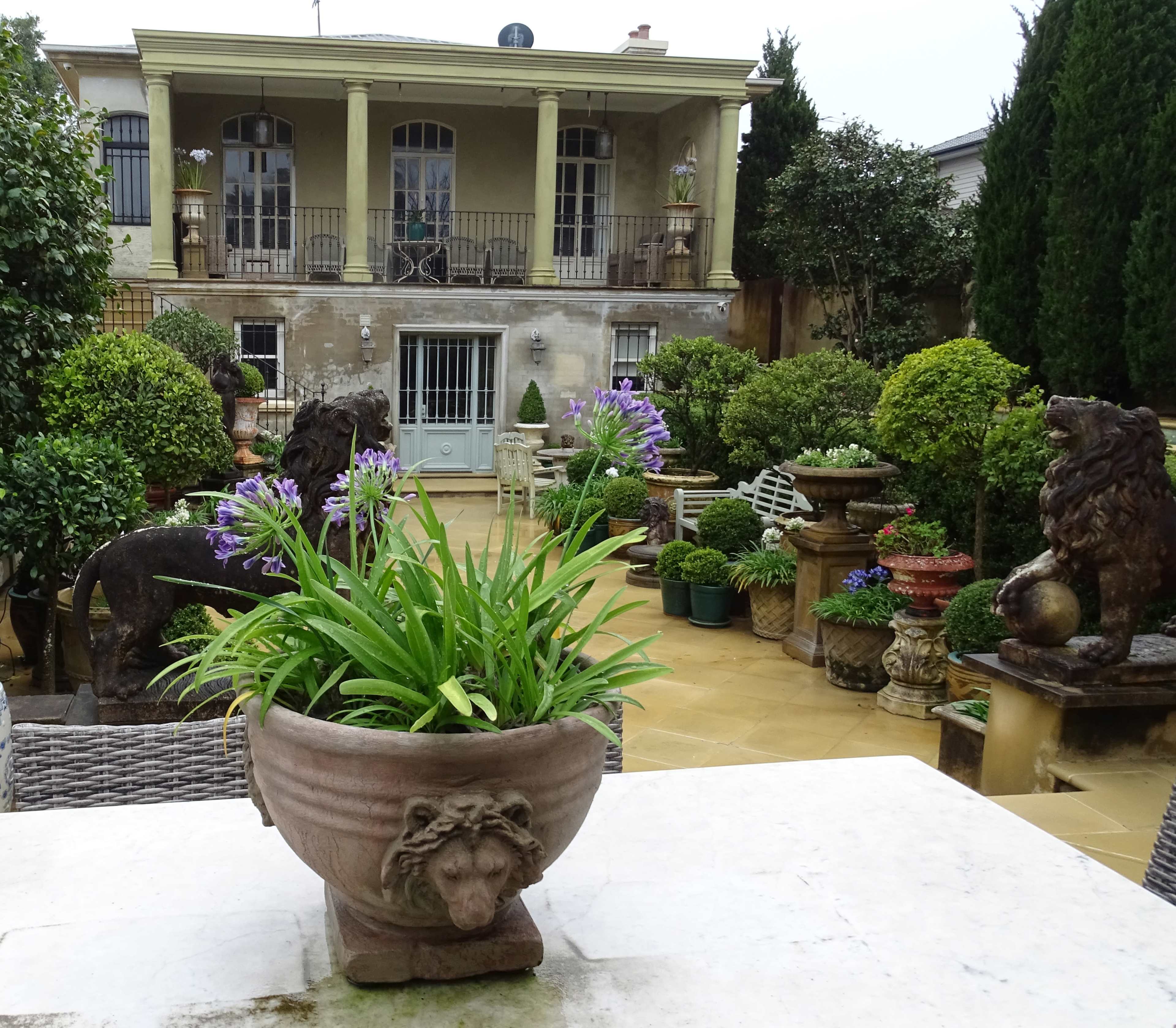A decorative garden features a stone lion planter in the foreground, lush greenery, and a grand house with balconies in the background.