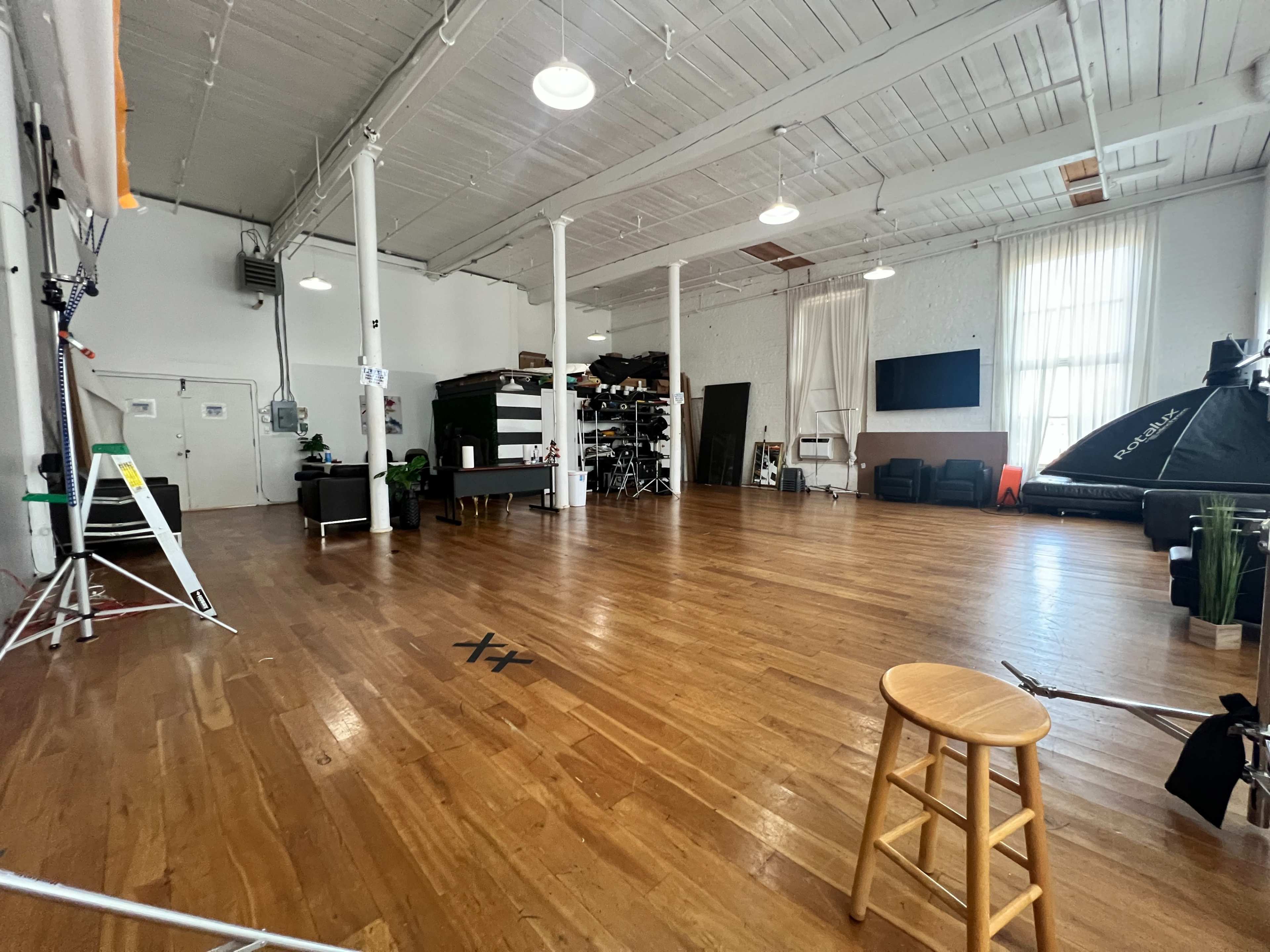 The image shows a spacious, well-lit studio with hardwood floors, tall ceilings, and various photography equipment arranged neatly along the walls.