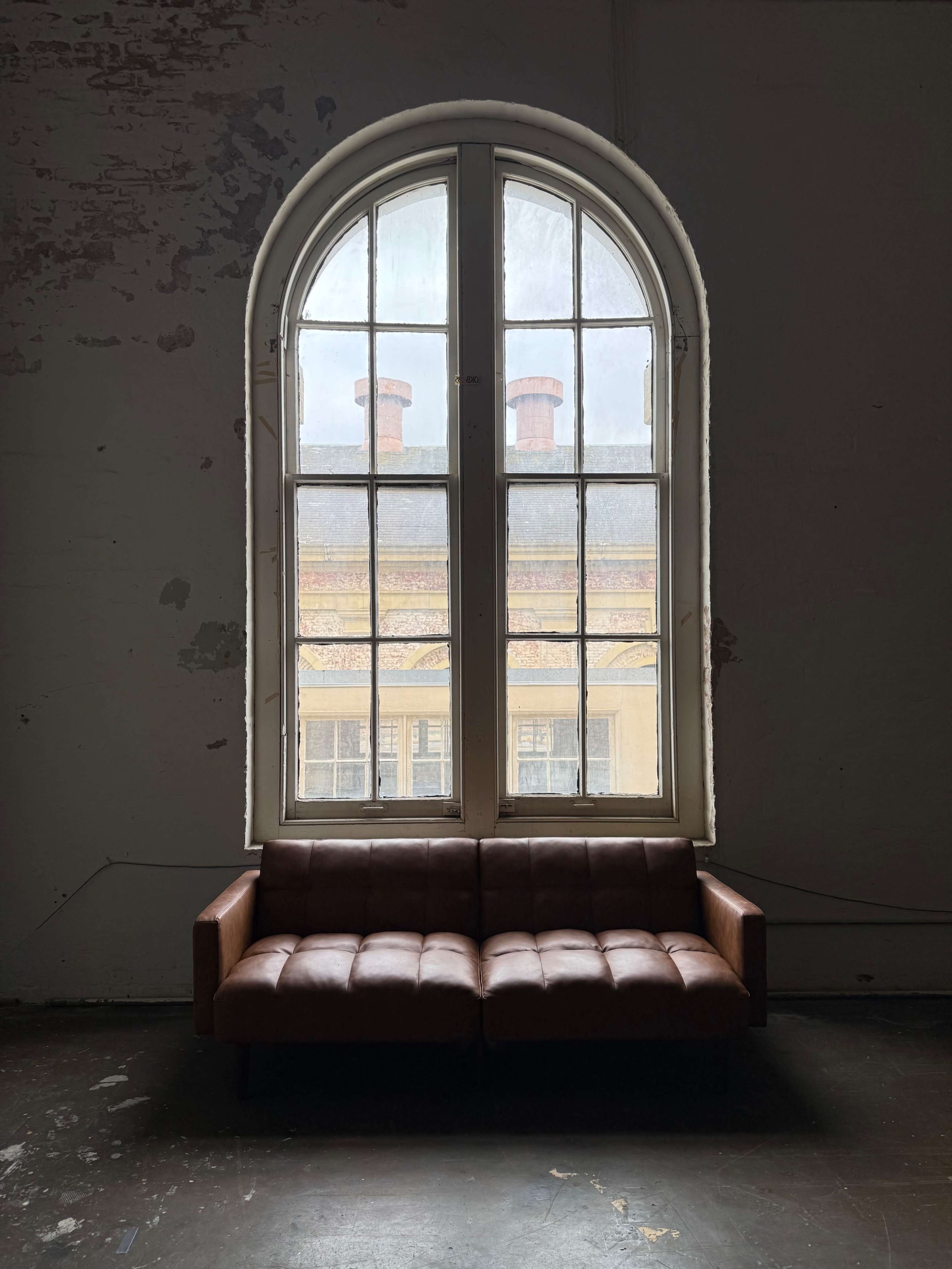 A brown leather sofa sits in front of a large arched window with multiple panes, revealing a view of red-brick chimneys outside.