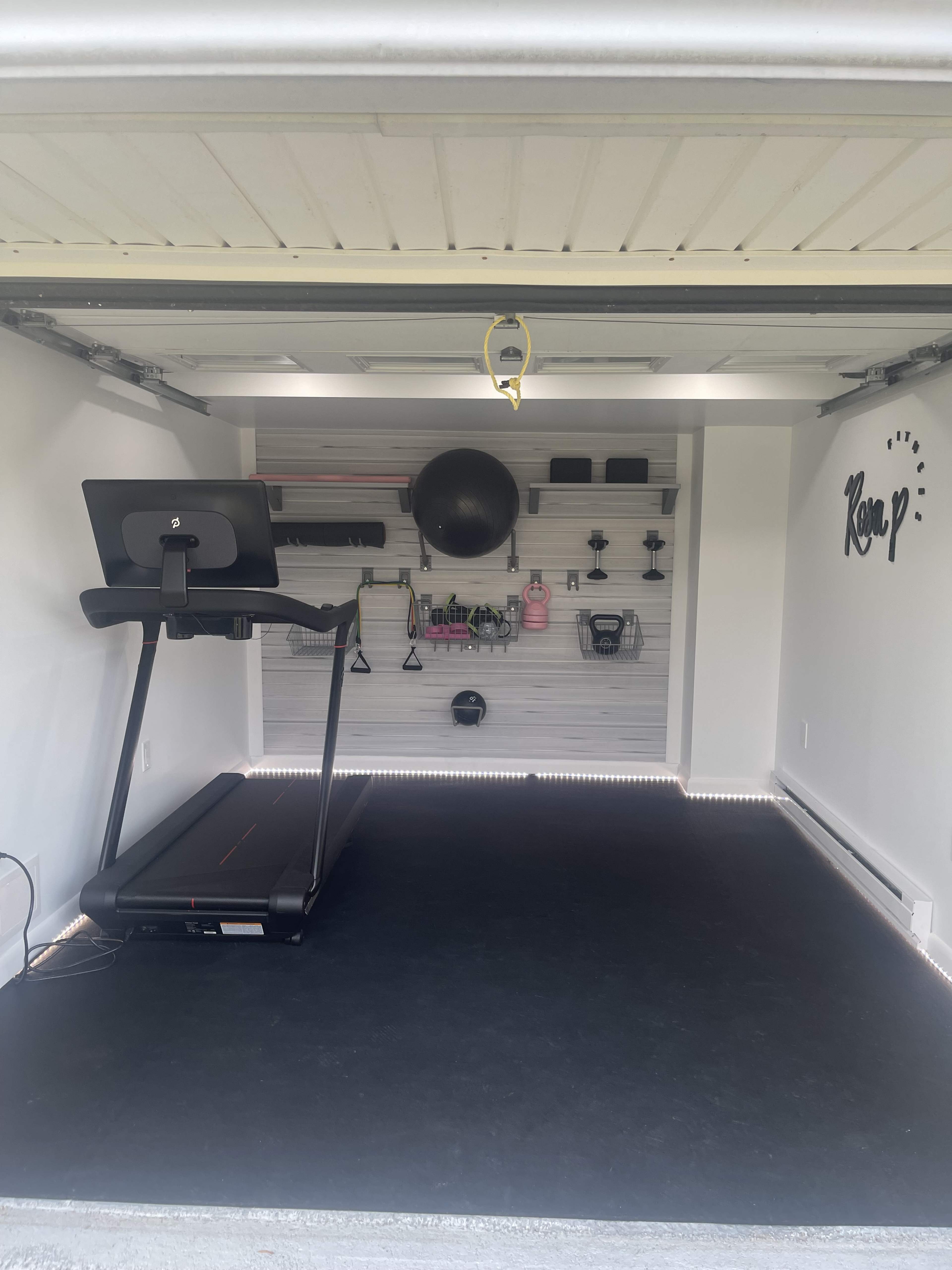 A neatly organized home gym inside a garage, featuring a treadmill, exercise equipment, and a workout ball on a wall-mounted storage system.