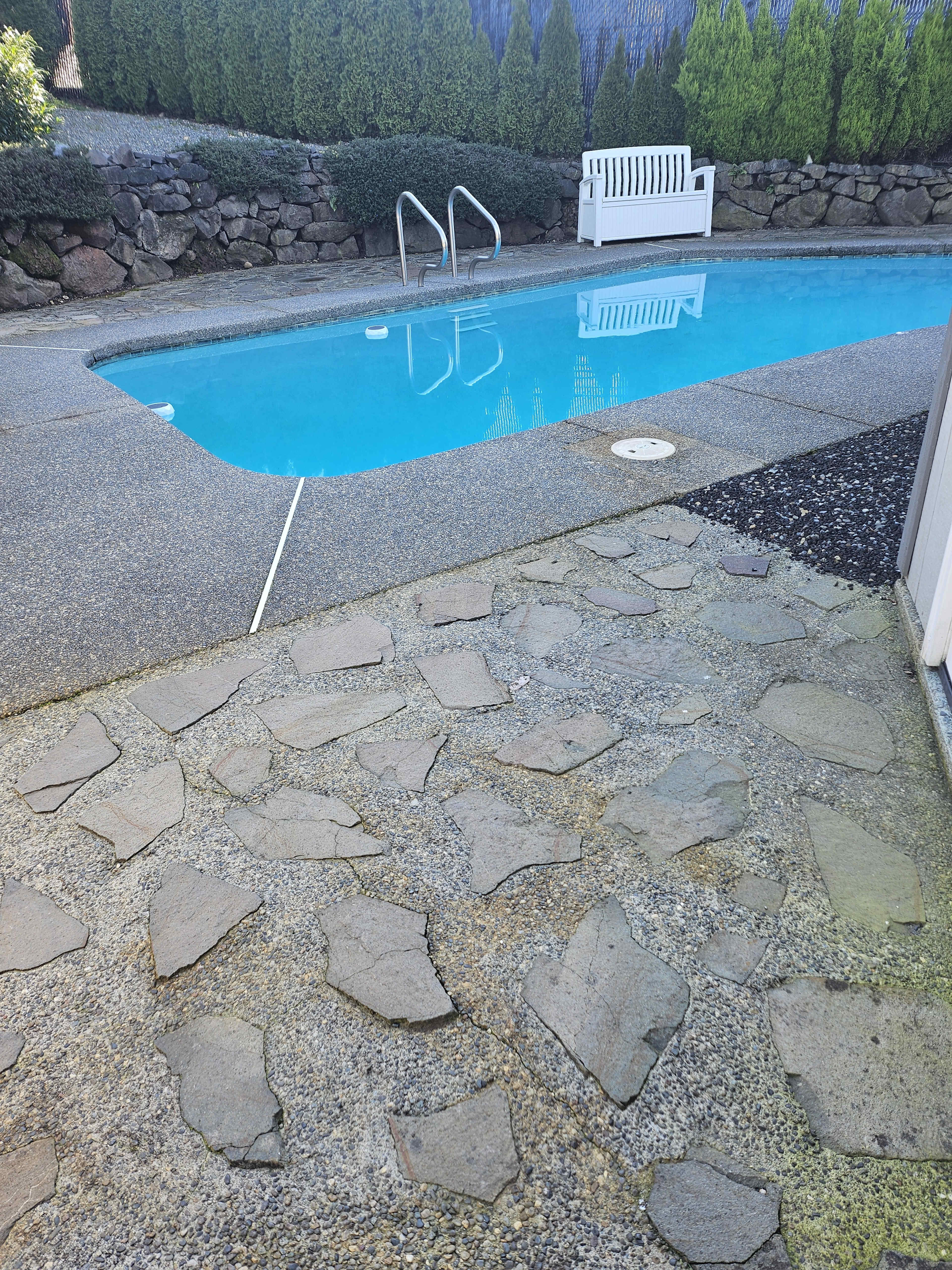 The image shows a swimming pool surrounded by a stone patio, with a white bench and green shrubbery in the background.