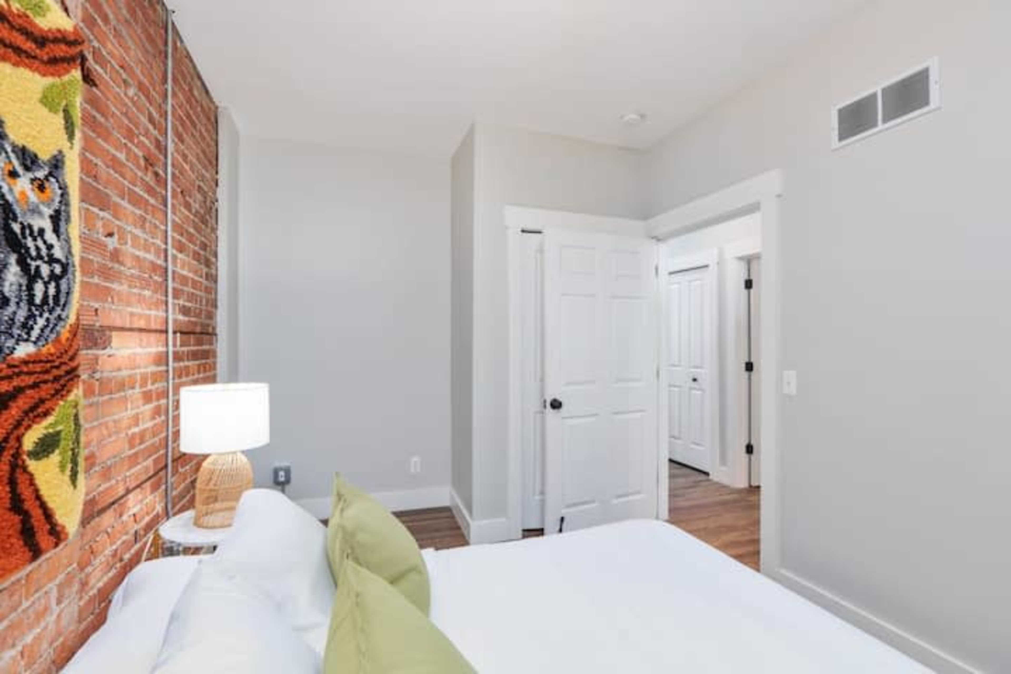 A bedroom with a bed, a bedside lamp, exposed brick wall, and a door leading to a hallway.