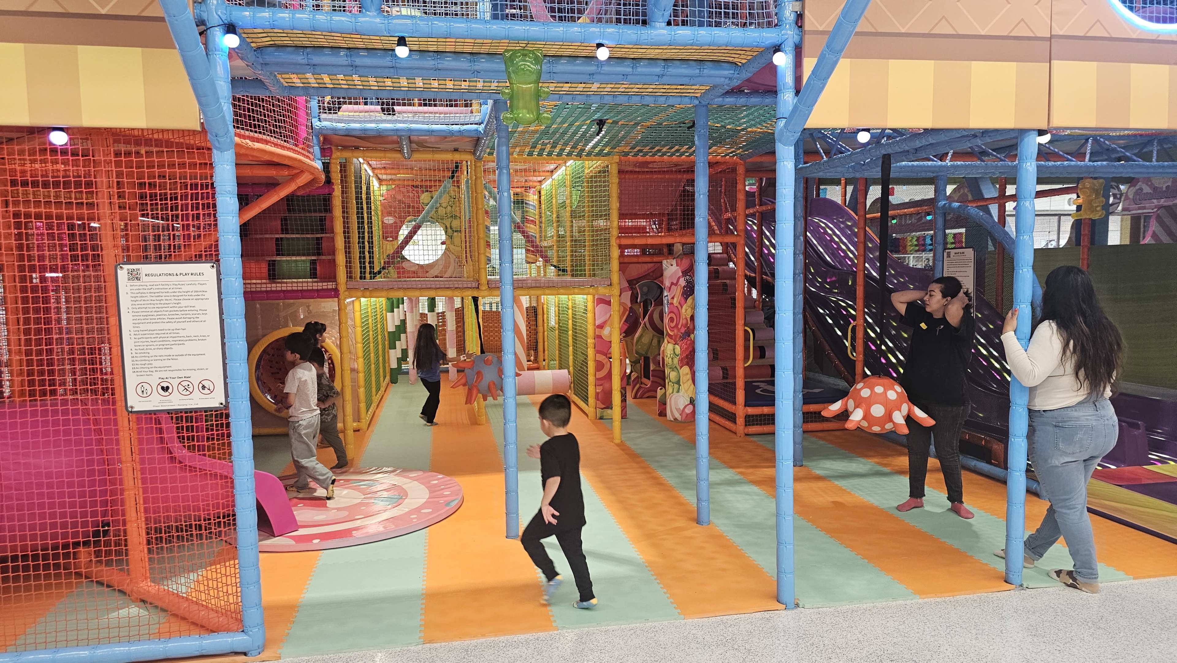 The image shows an indoor play area with colorful structures, children playing in various sections, and adults watching along the sides.