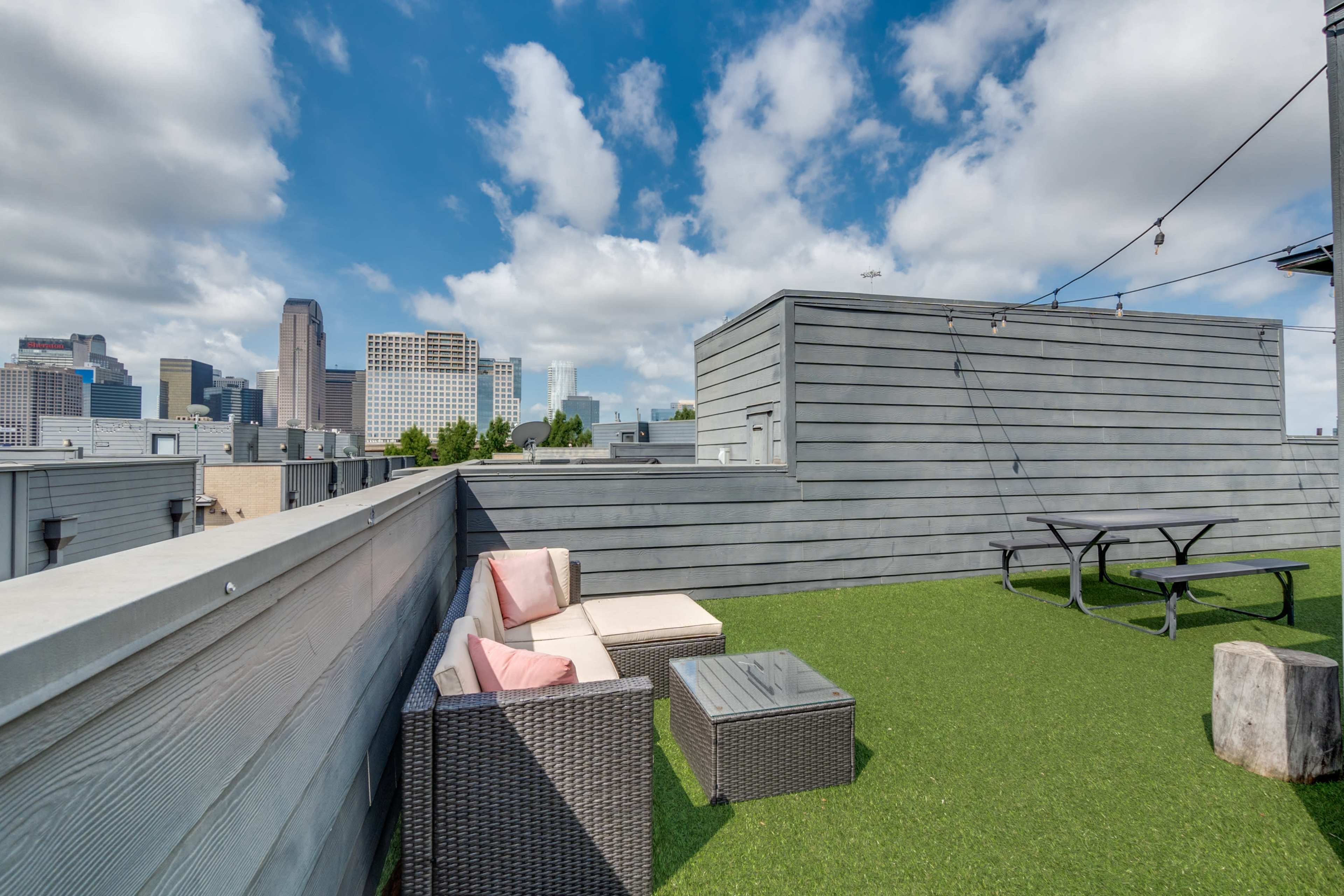 A rooftop space with artificial grass features seating made of wicker and cushions, alongside a small table, with a city skyline visible in the background under partly cloudy skies.