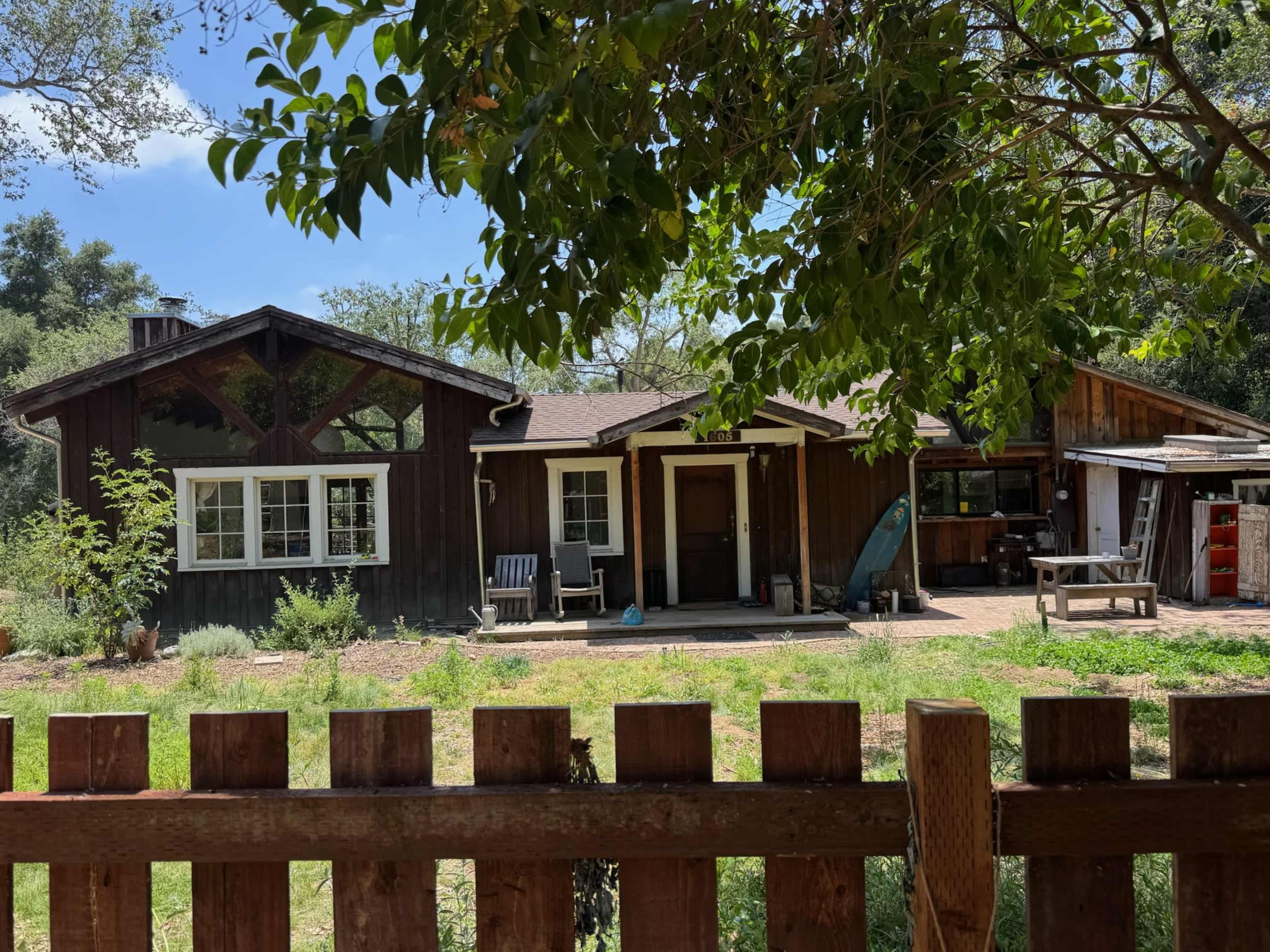 A wooden house with a porch, surrounded by greenery, is visible behind a wooden fence.