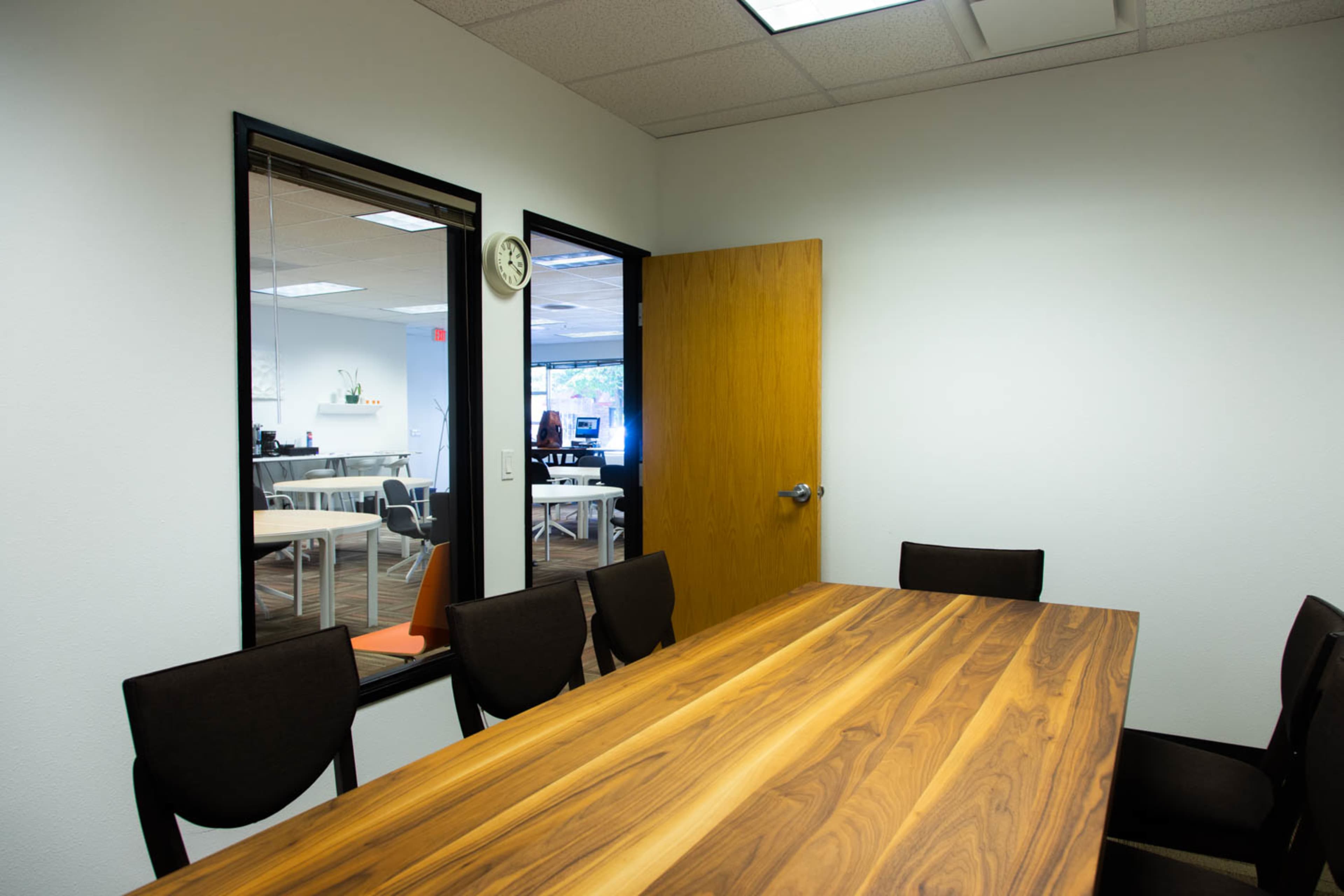A conference room with a wooden table and a view through a glass wall into an adjacent office area.