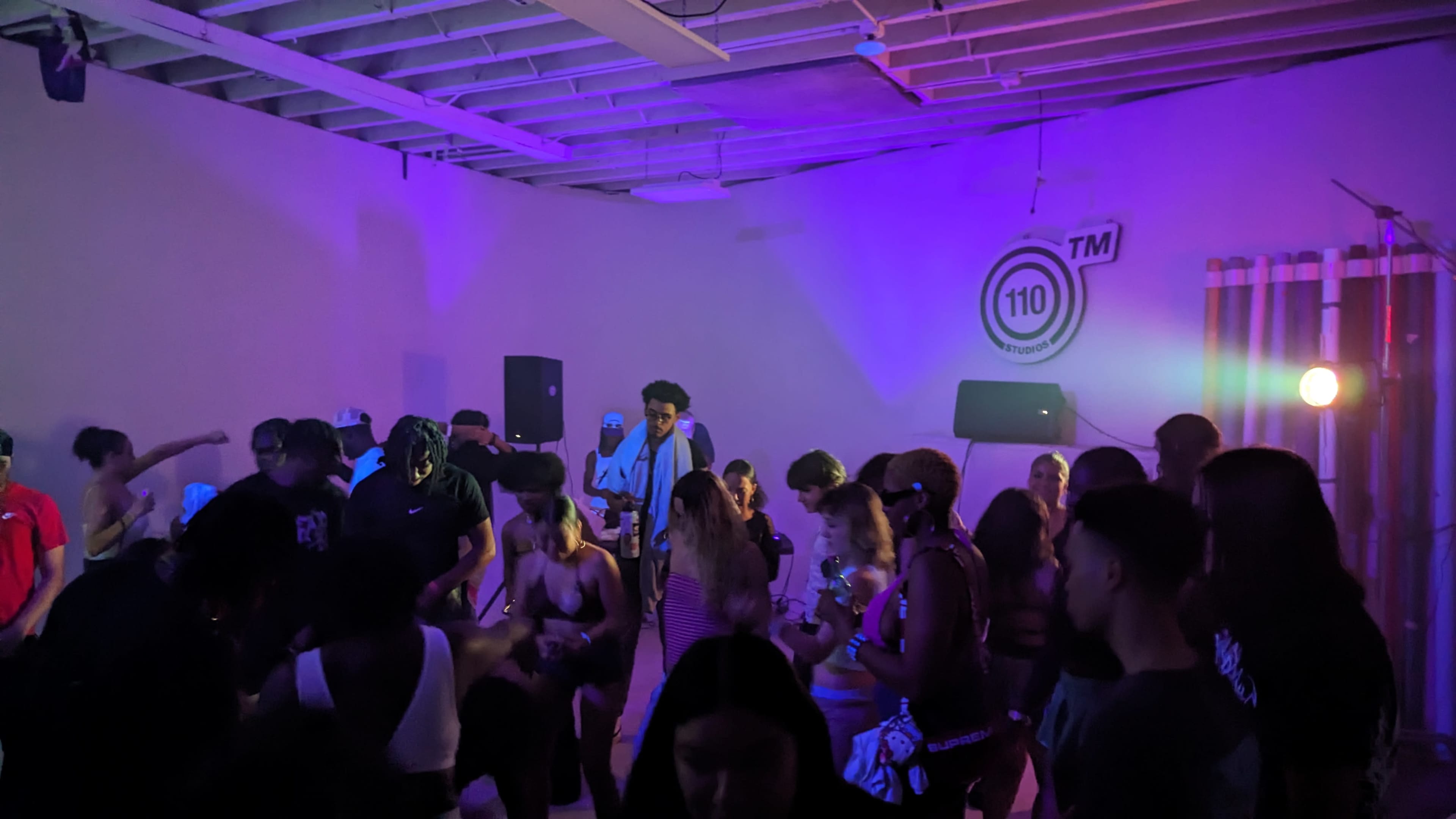 A crowd of people dances in a dimly lit room with purple lighting and a large speaker setup.