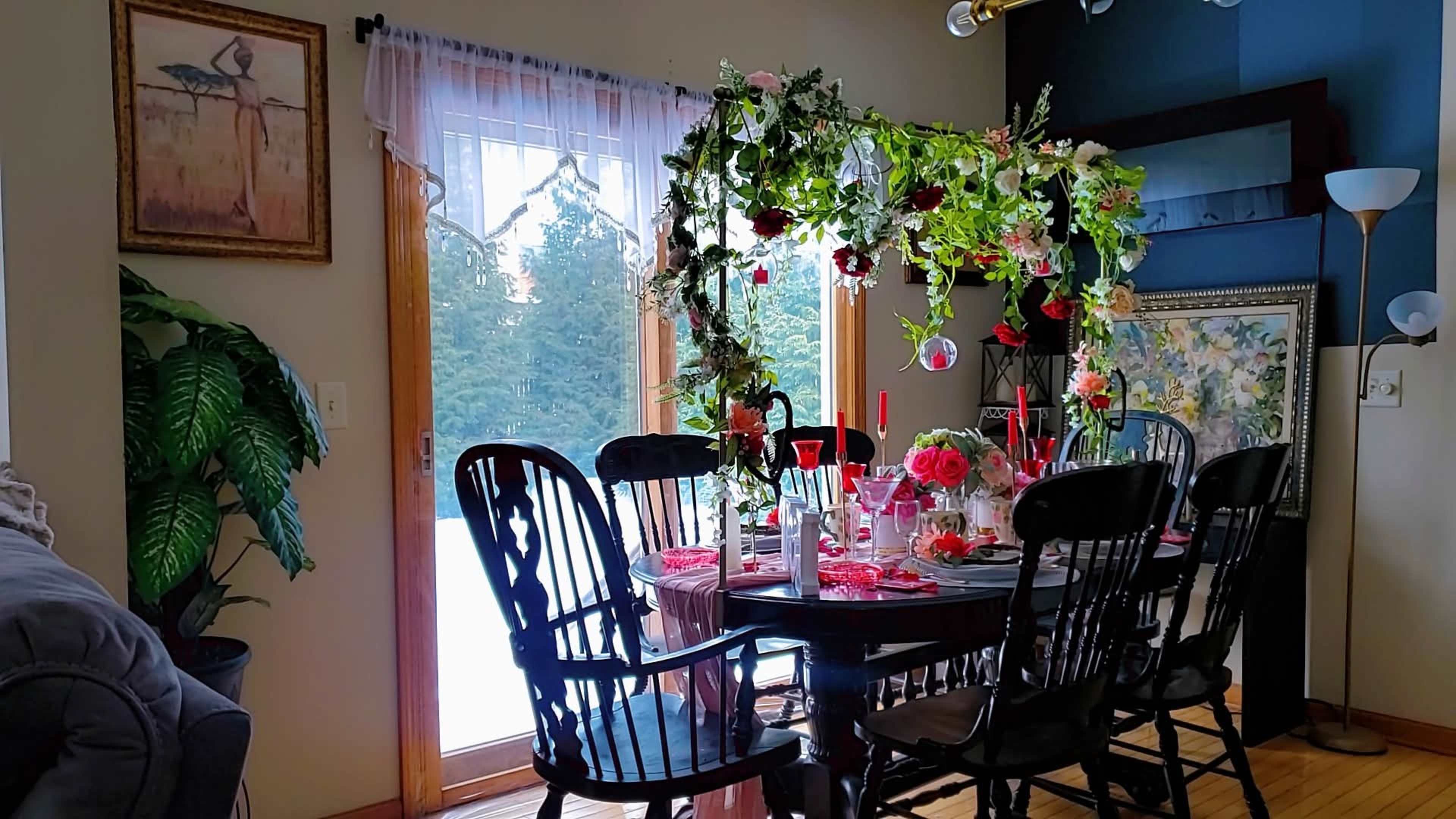 A dining area features a table set for a meal, surrounded by black wooden chairs and adorned with floral decorations, while sunlight streams through a large window.