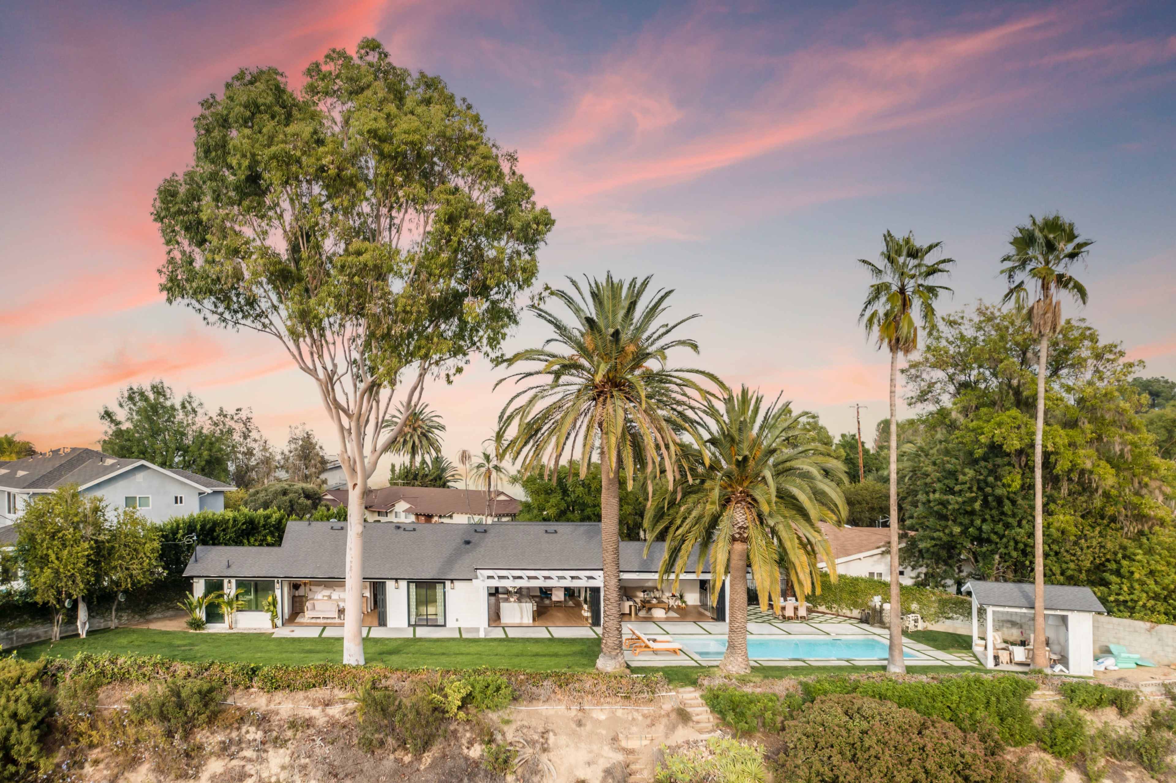 The image shows a modern single-story house with a swimming pool and palm trees, set against a colorful sunset sky.
