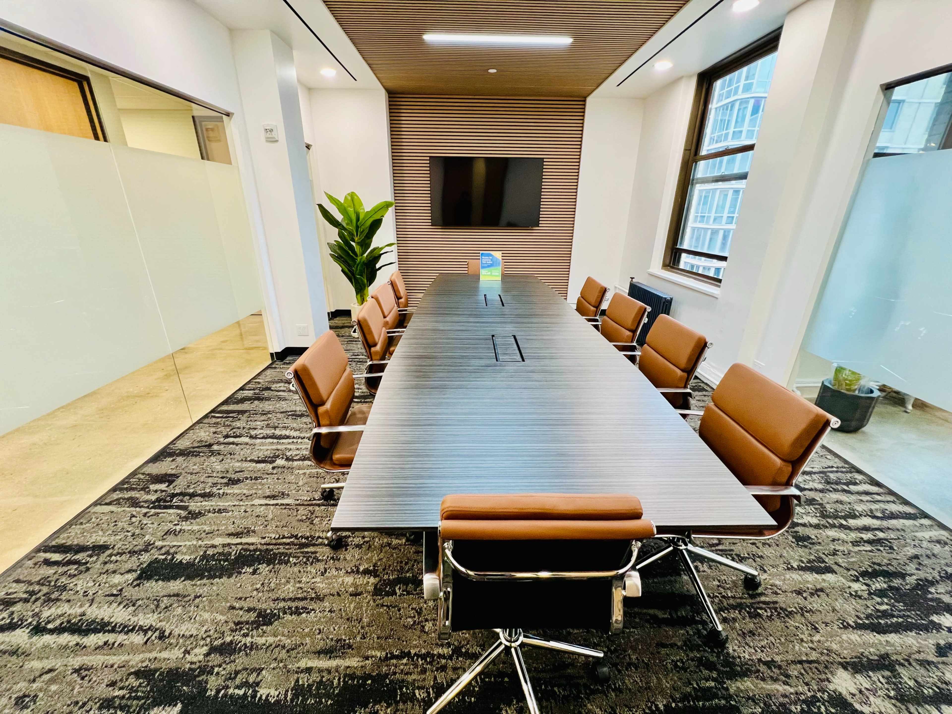 A large conference room features a long, rectangular wooden table surrounded by brown leather chairs, with a television mounted on the wall and a potted plant in the corner.
