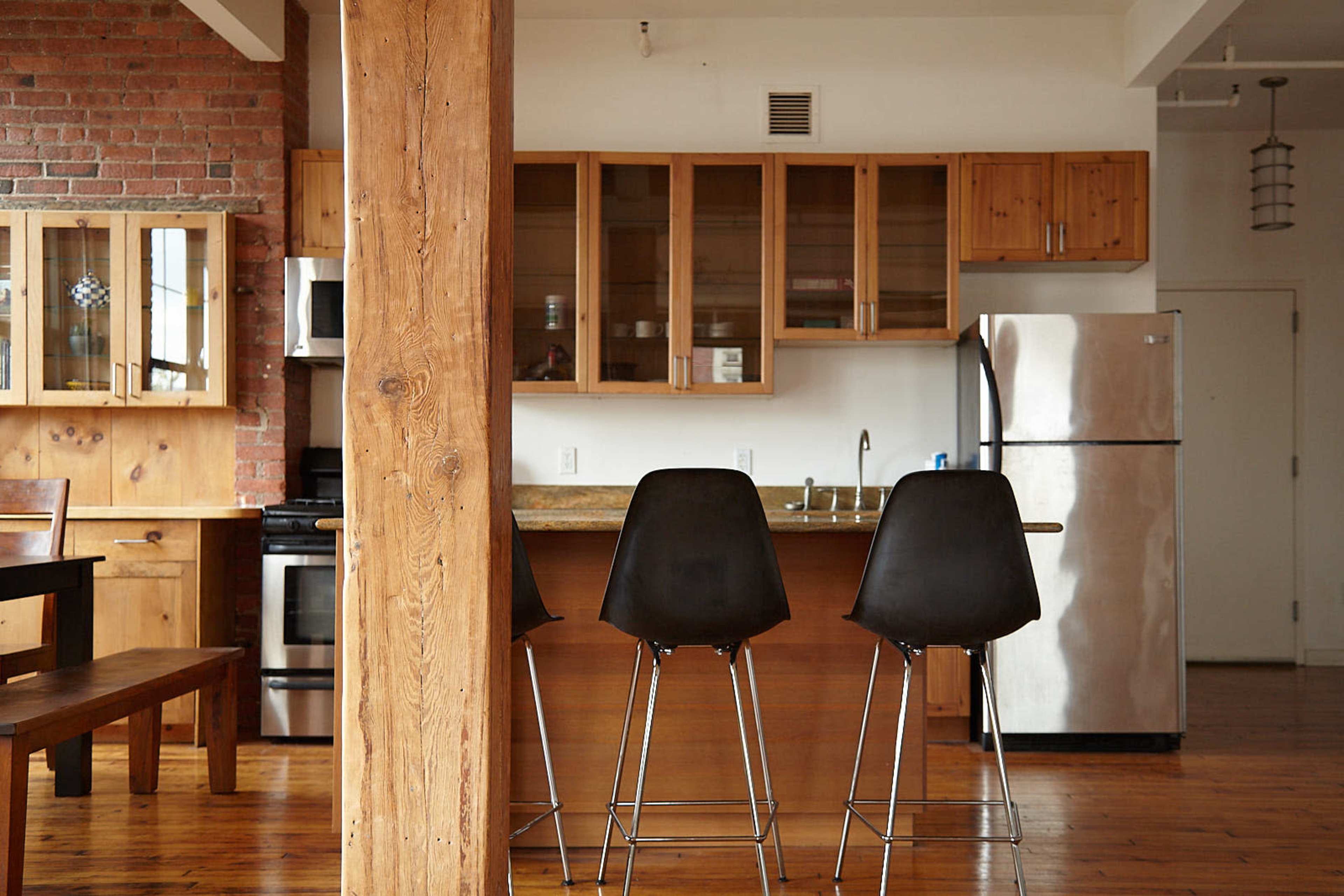 The image shows a modern kitchen with wooden cabinetry, a stainless steel refrigerator, and three black bar stools positioned at a counter.