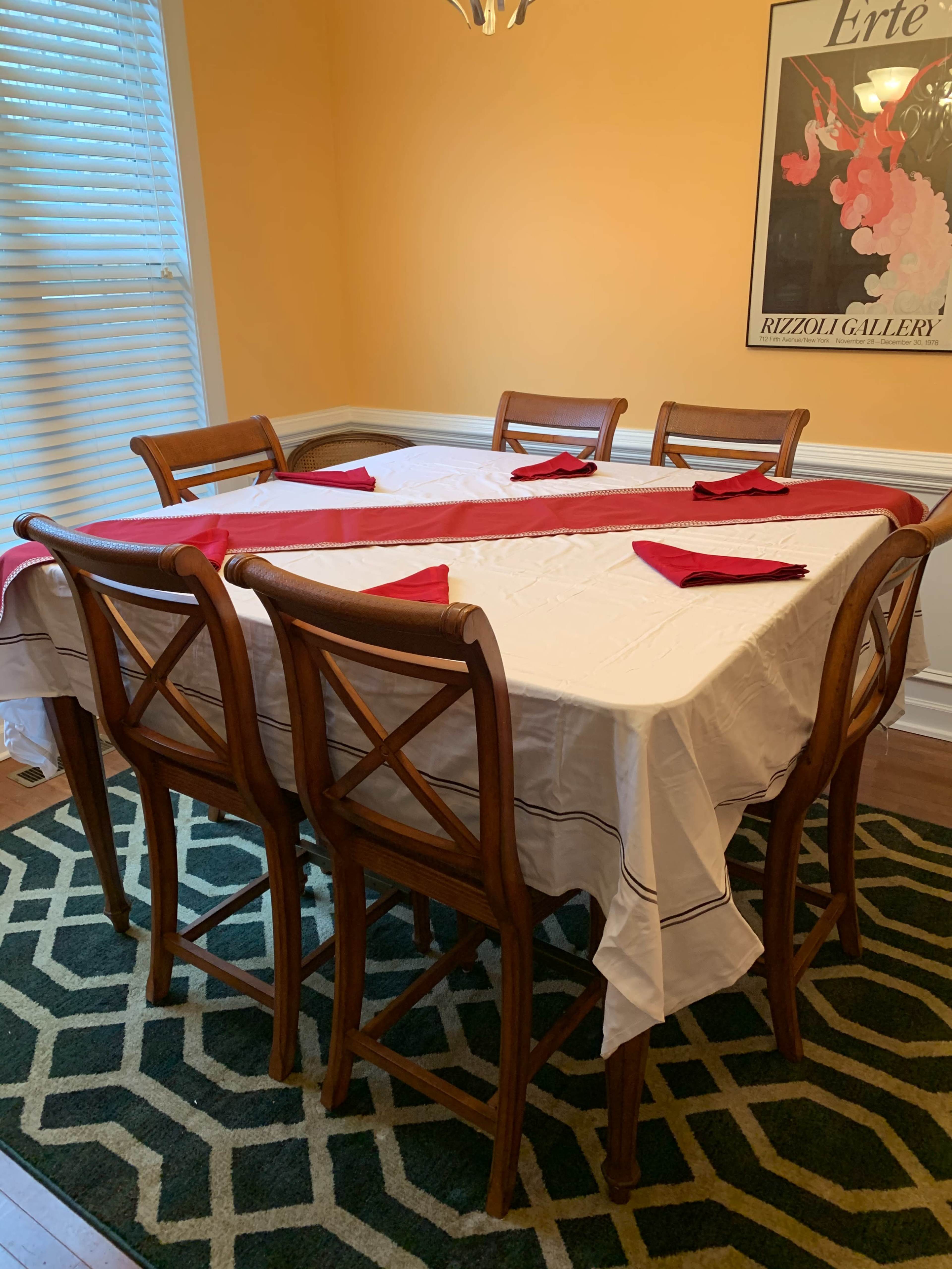 A rectangular dining table with a white tablecloth and red napkins is set in a well-lit room with yellow walls and a green patterned rug.