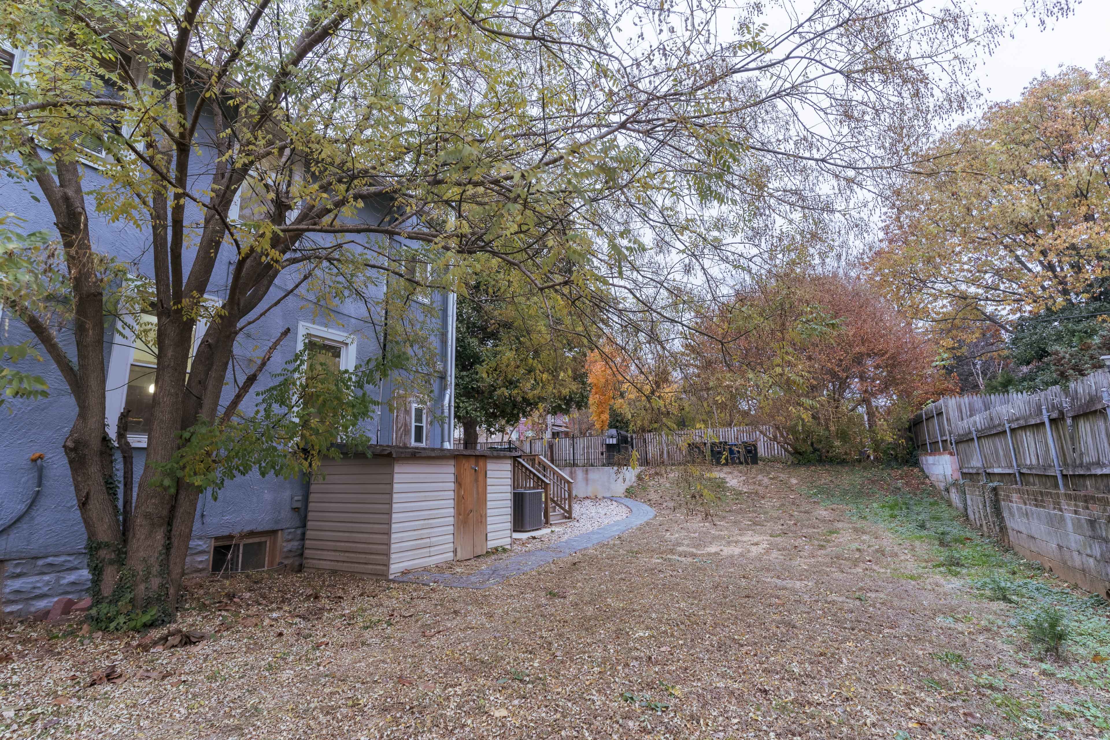 A backyard features a wooden shed next to a blue house, with trees and foliage in various autumn colors.