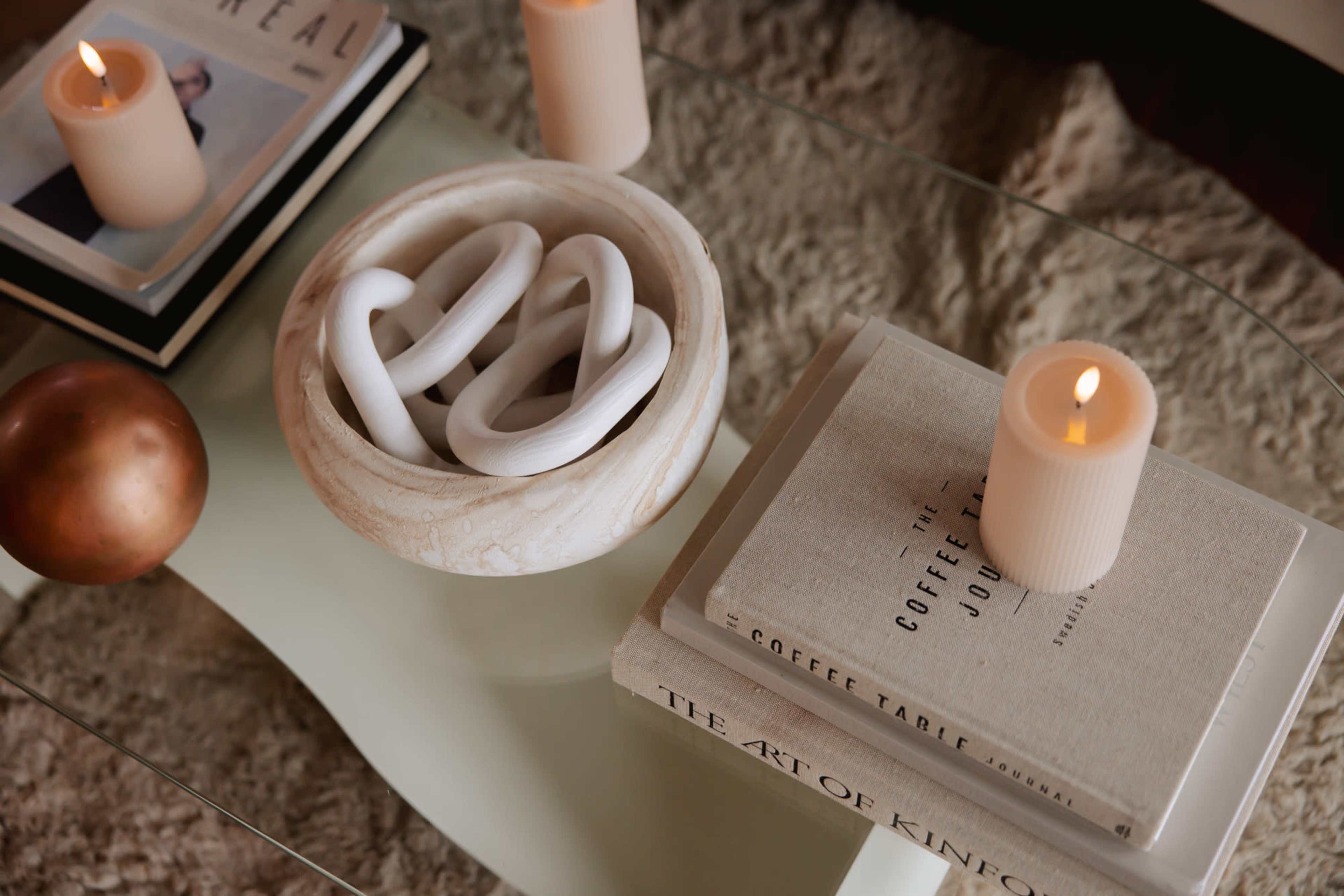 A coffee table displays stacked books, a circular stone bowl with white decorative objects, and lit candles.