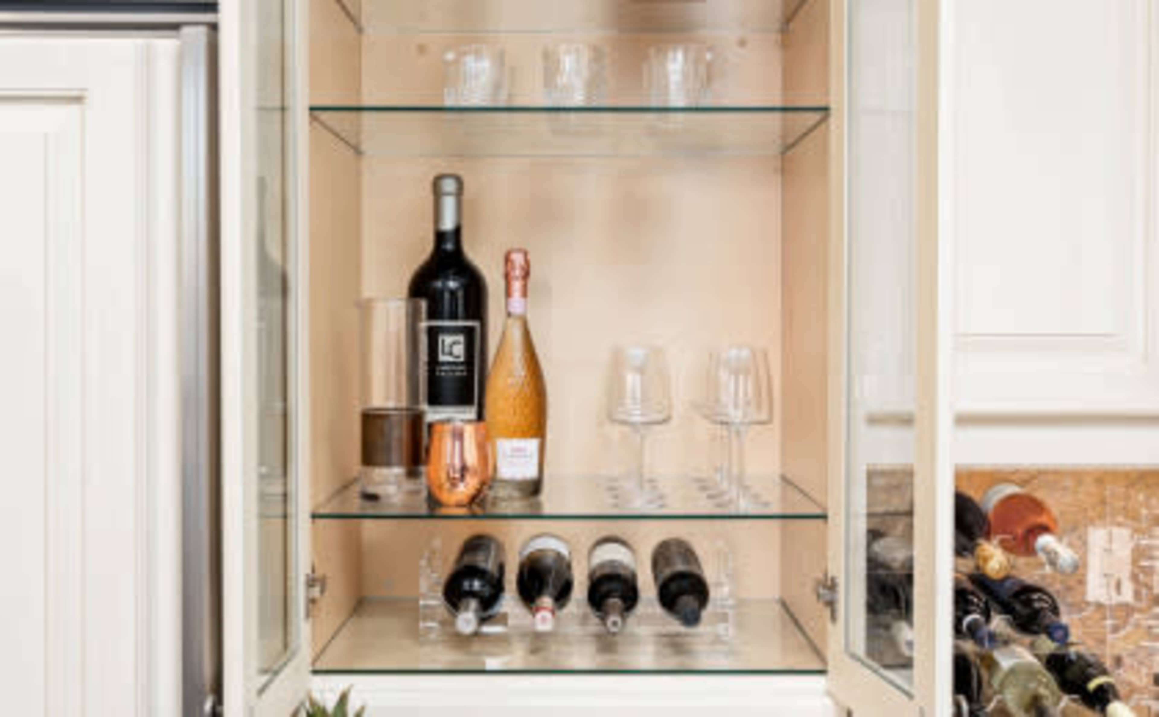 A glass cabinet containing various bottles of wine, champagne, and several drinking glasses organized on shelves.