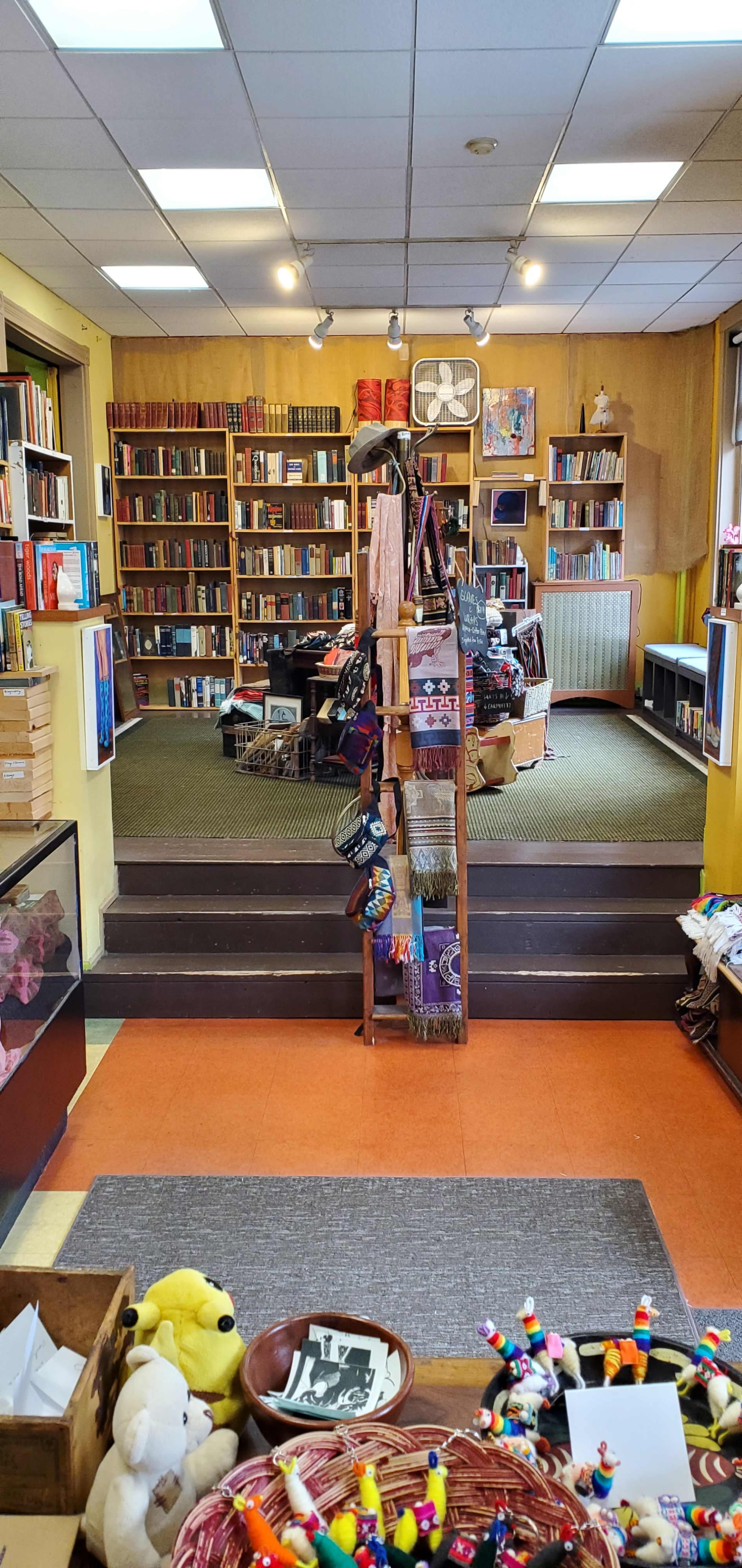 A cozy bookstore with wooden shelves filled with books, a display of handmade crafts in the foreground, and a staircase leading to an upper level.