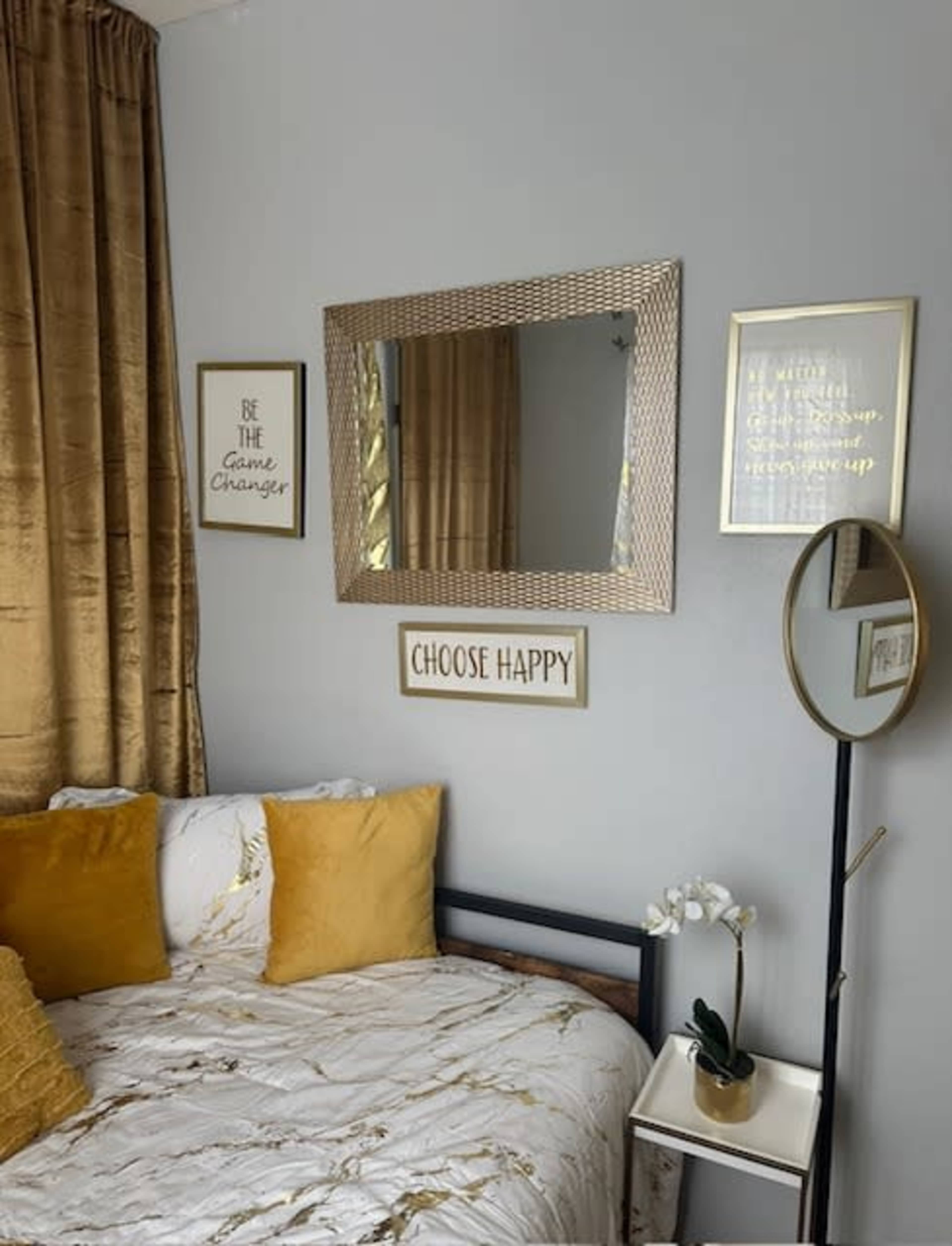 A cozy bedroom with a bed featuring yellow and white bedding, a mirror on the wall, and decorative signs above the bed, accompanied by a side table and a standing mirror.