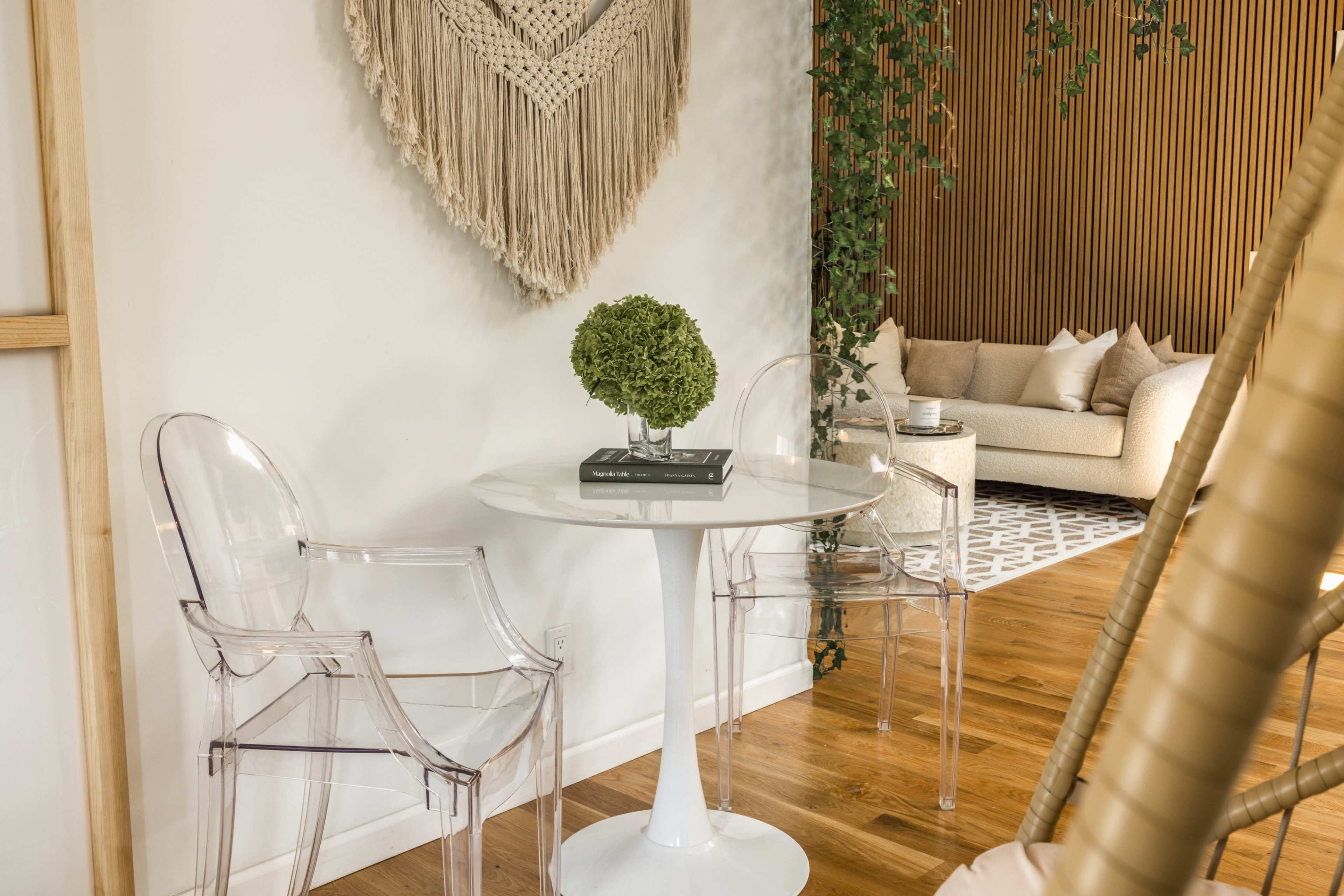 A small dining area features two clear chairs around a round white table, with a decorative wall hanging and a cozy sofa in the background.