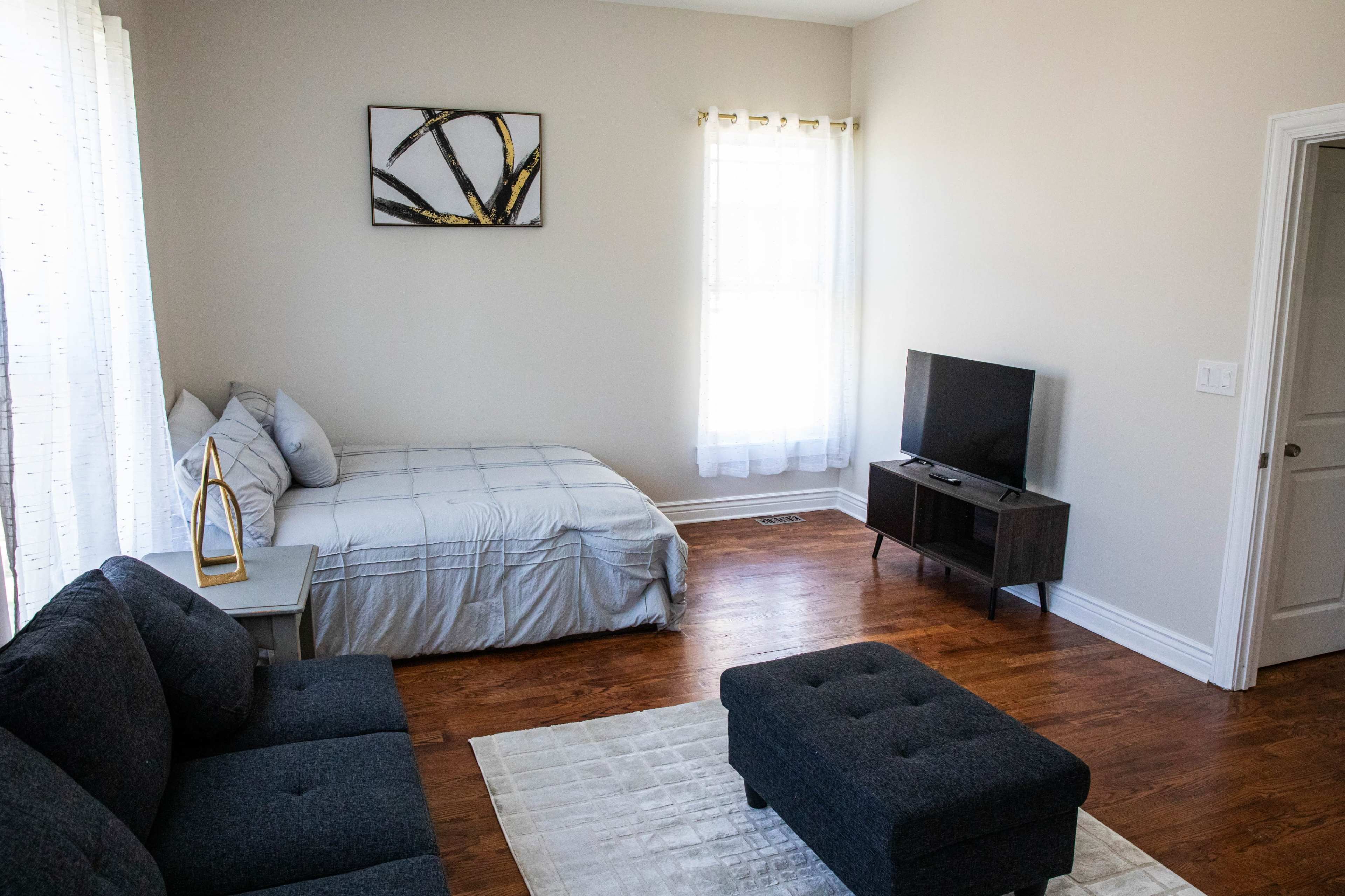 The image shows a furnished living space with a bed, a couch, a coffee table, a television, and large windows allowing natural light.