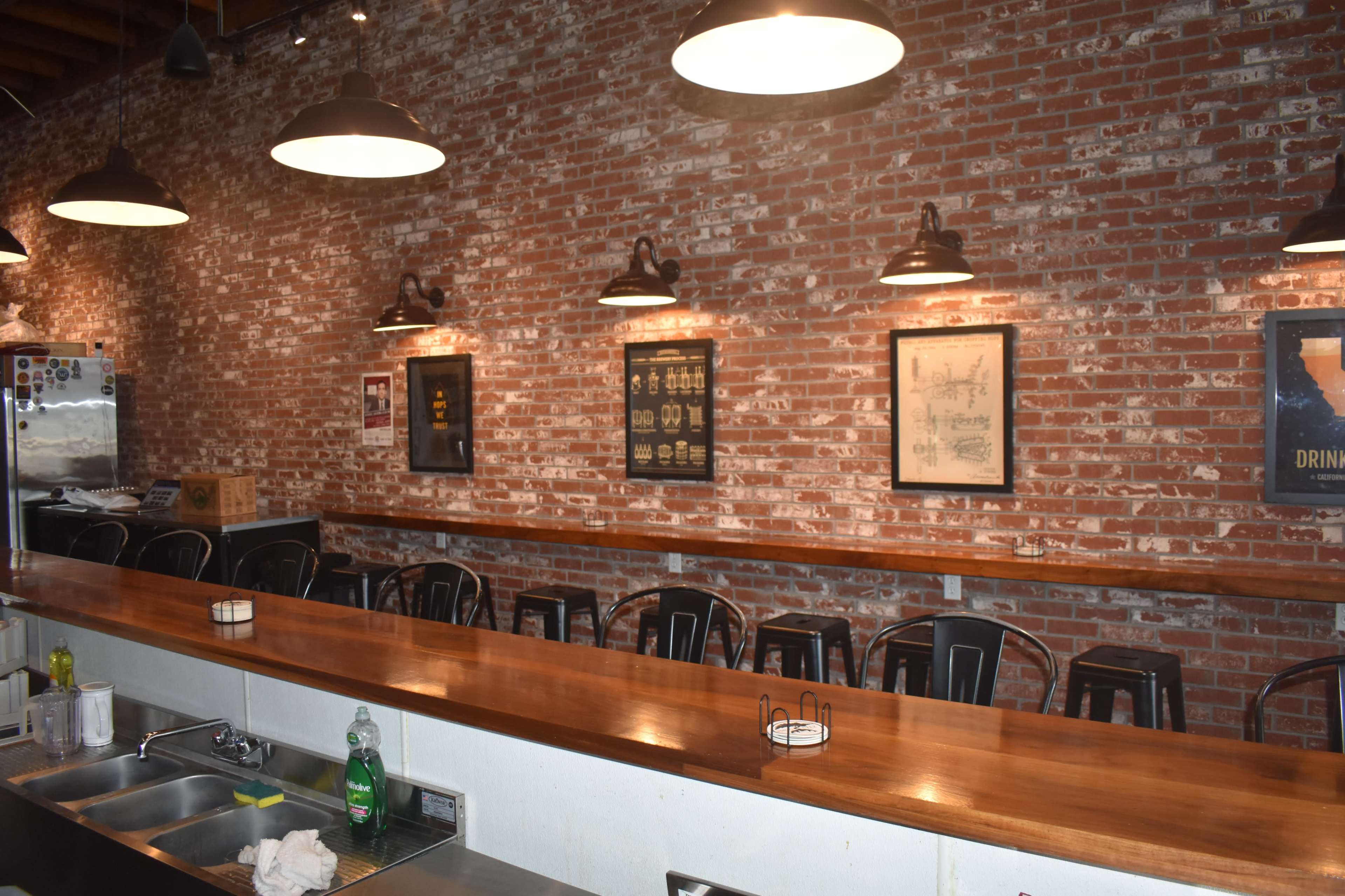 The image shows a restaurant interior with a long wooden bar, high stools, and brick walls adorned with framed artwork.