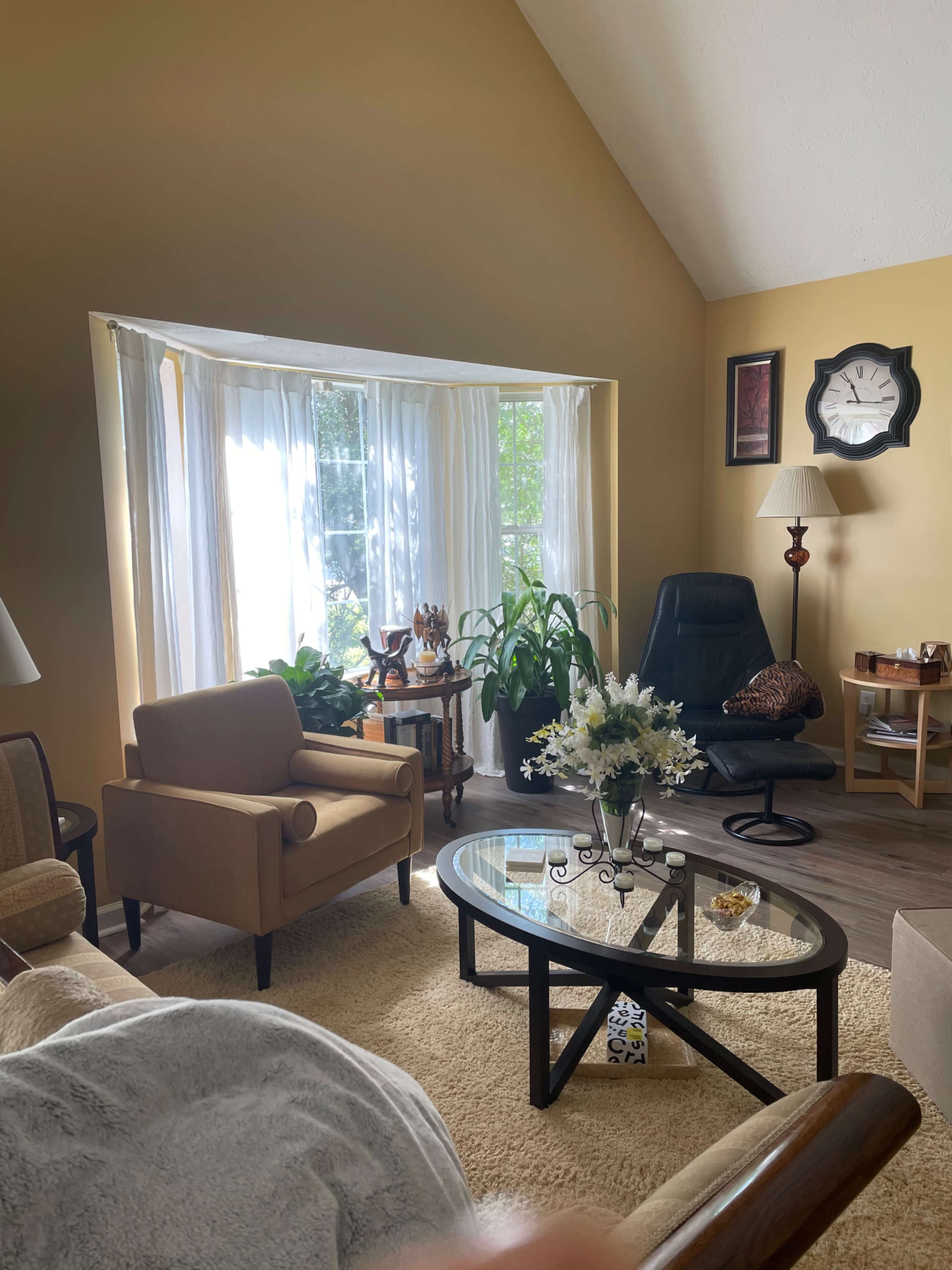 A cozy living room features a large window with white curtains, two armchairs, a coffee table with flowers, and a corner lamp.