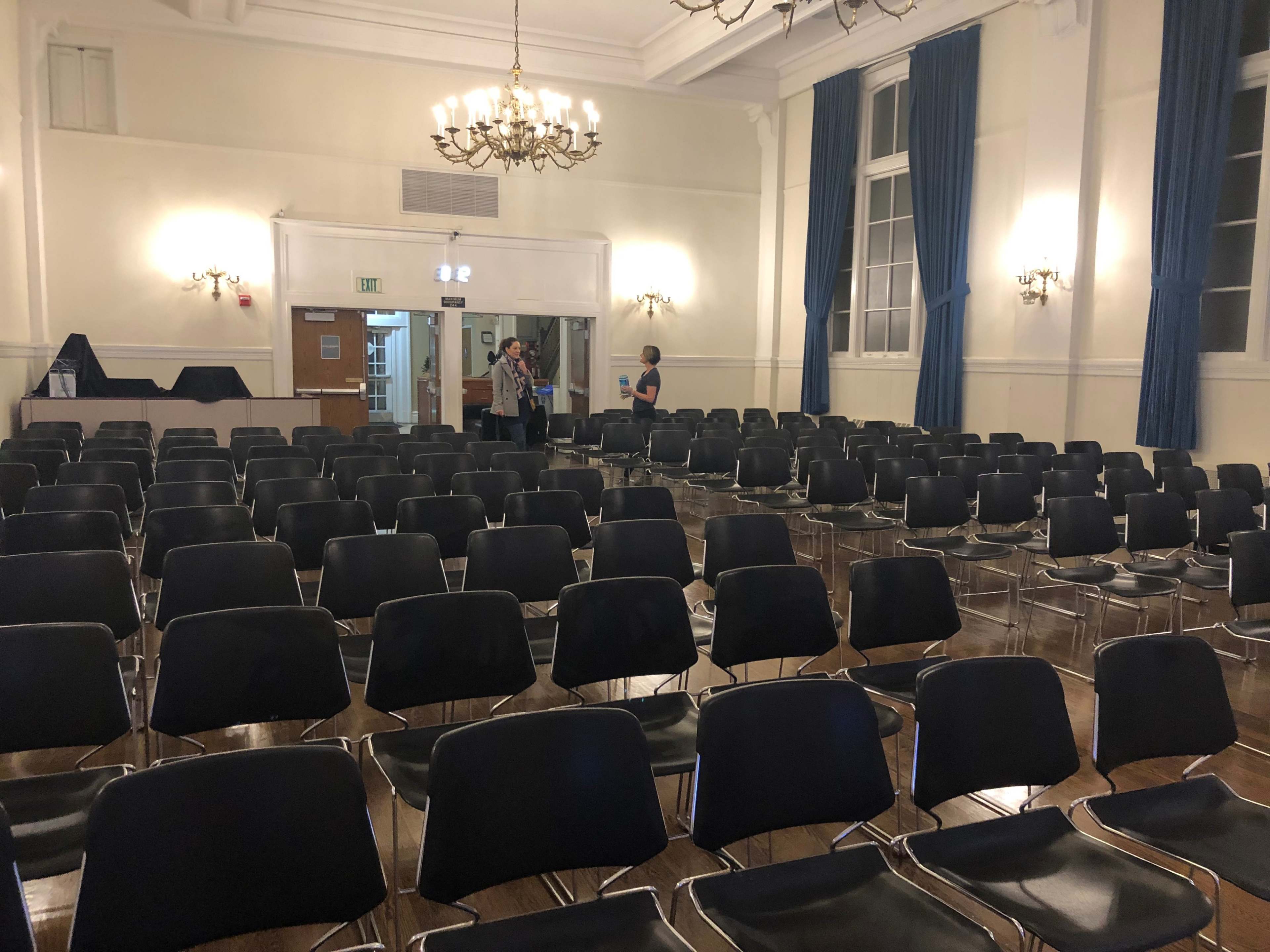 A large, empty room filled with rows of black chairs, with a few people standing in the background near the entrance.