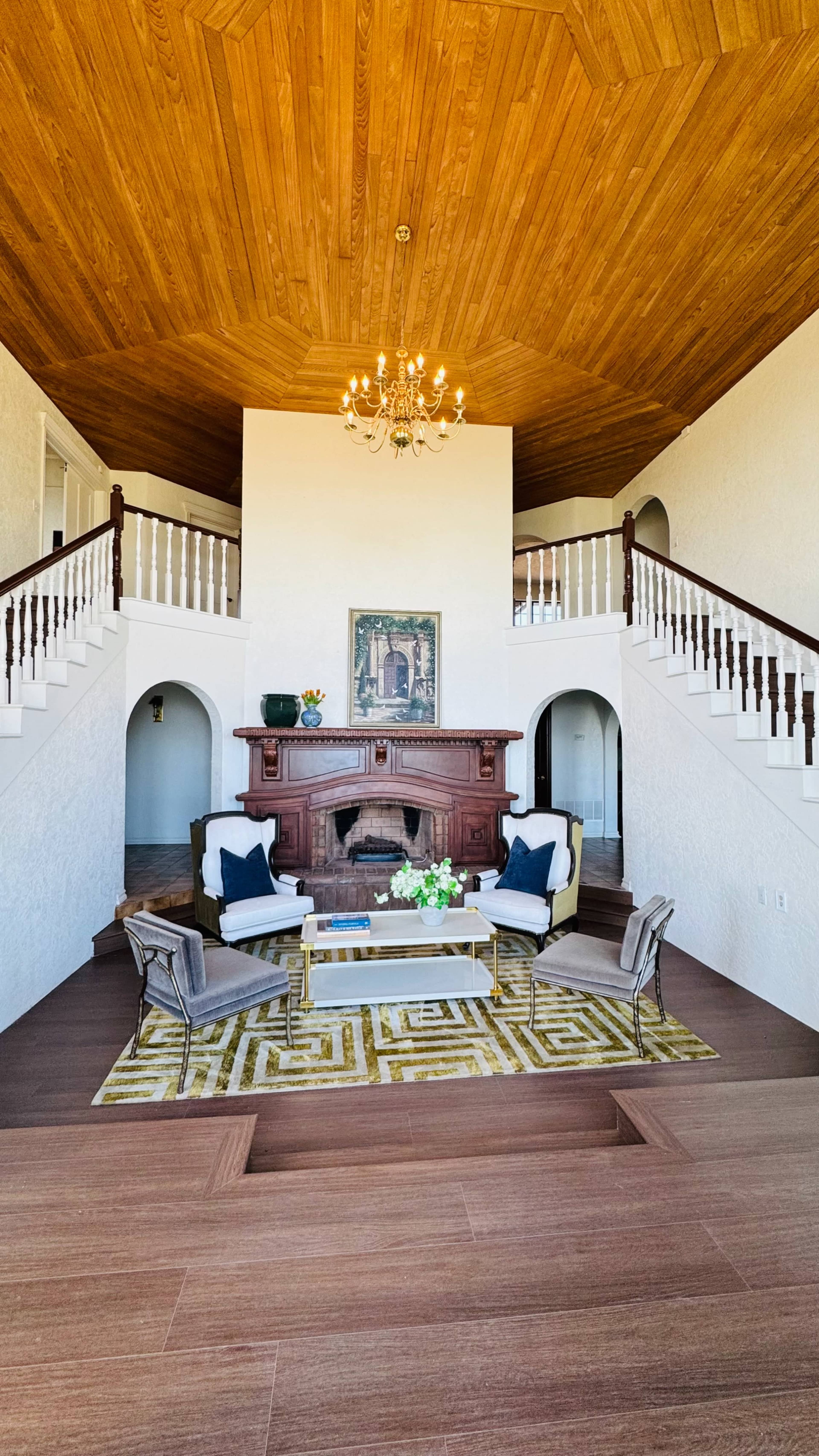 A spacious living room features a central seating area with a coffee table and decorative rug, framed by a high wooden ceiling and an elegant chandelier.