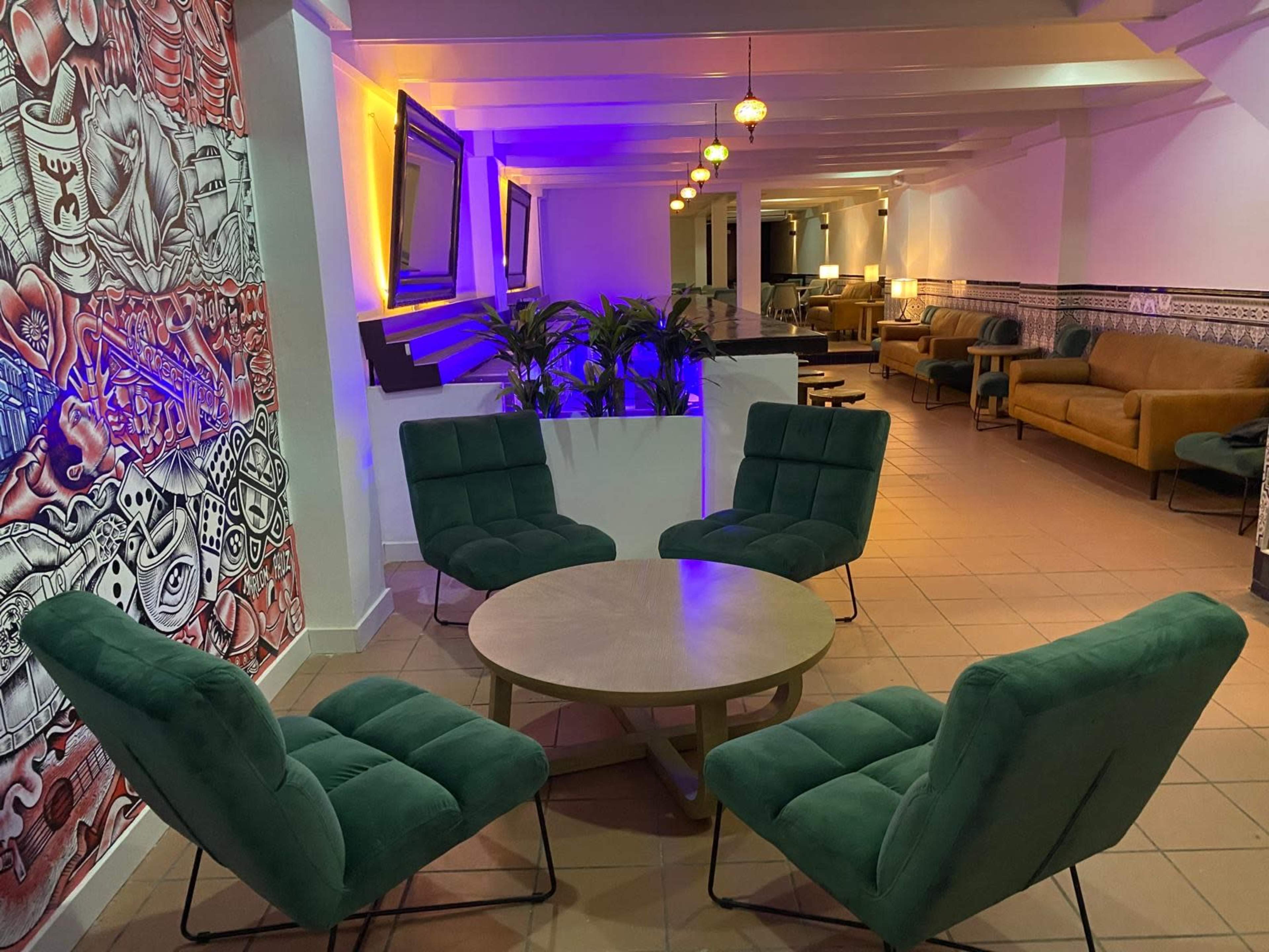 The image shows a lounge area with a circular table surrounded by four green upholstered chairs, colorful lighting, and a decorative wall mural.
