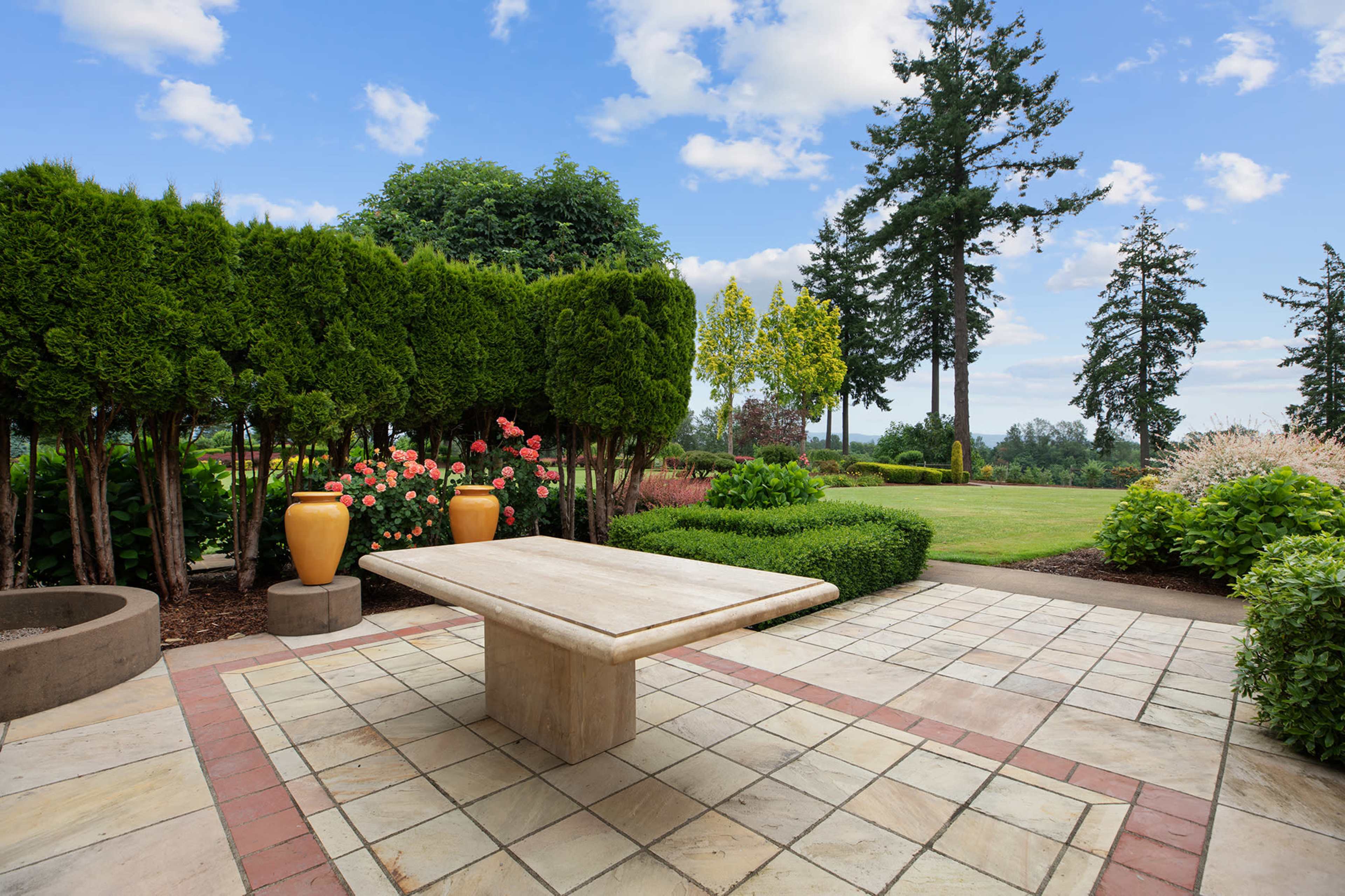 A landscaped outdoor area features a stone table surrounded by greenery and floral arrangements under a blue sky.
