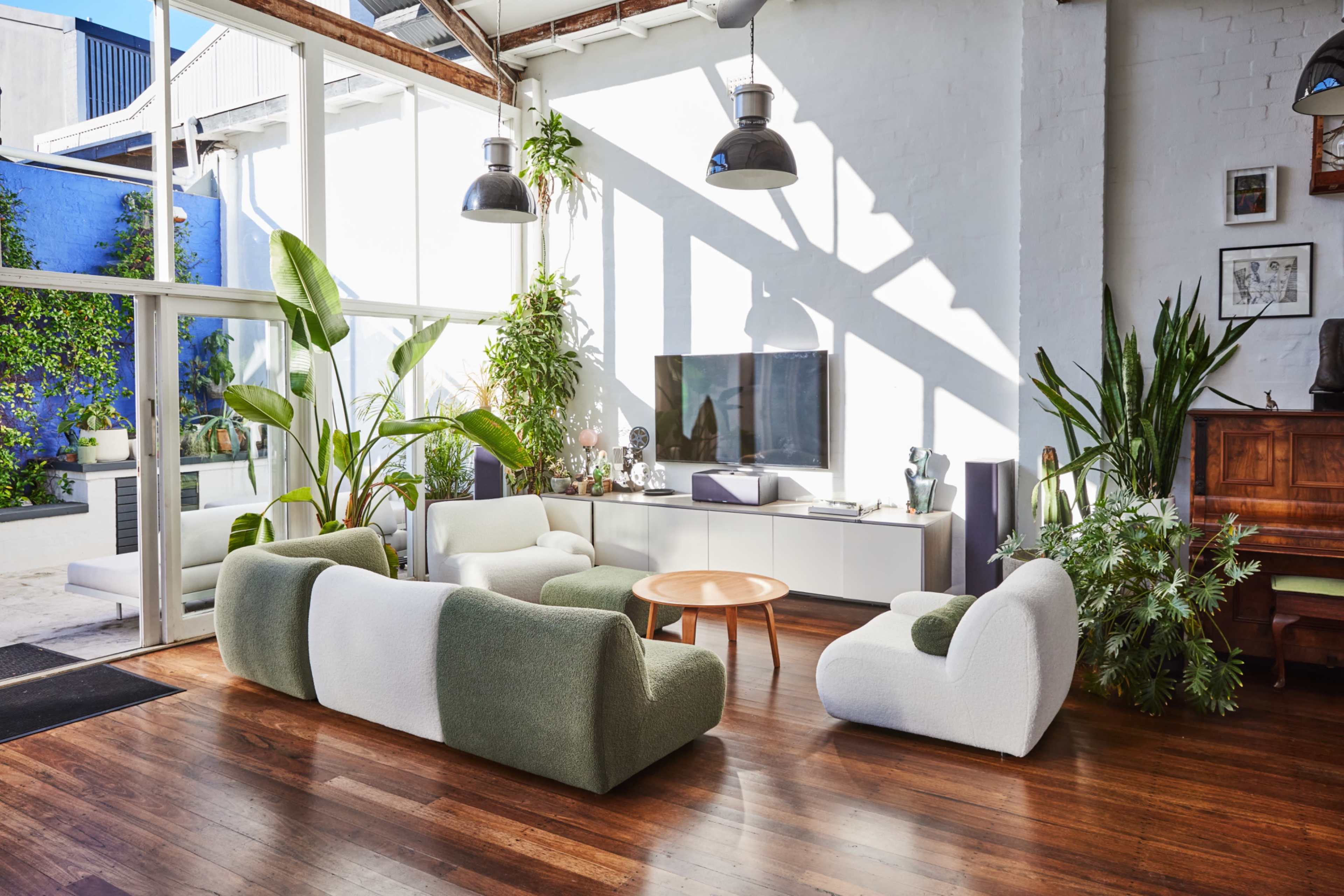 The image shows a modern living room with a mix of green and white curved sofas, a wooden coffee table, and large windows allowing natural light to fill the space.