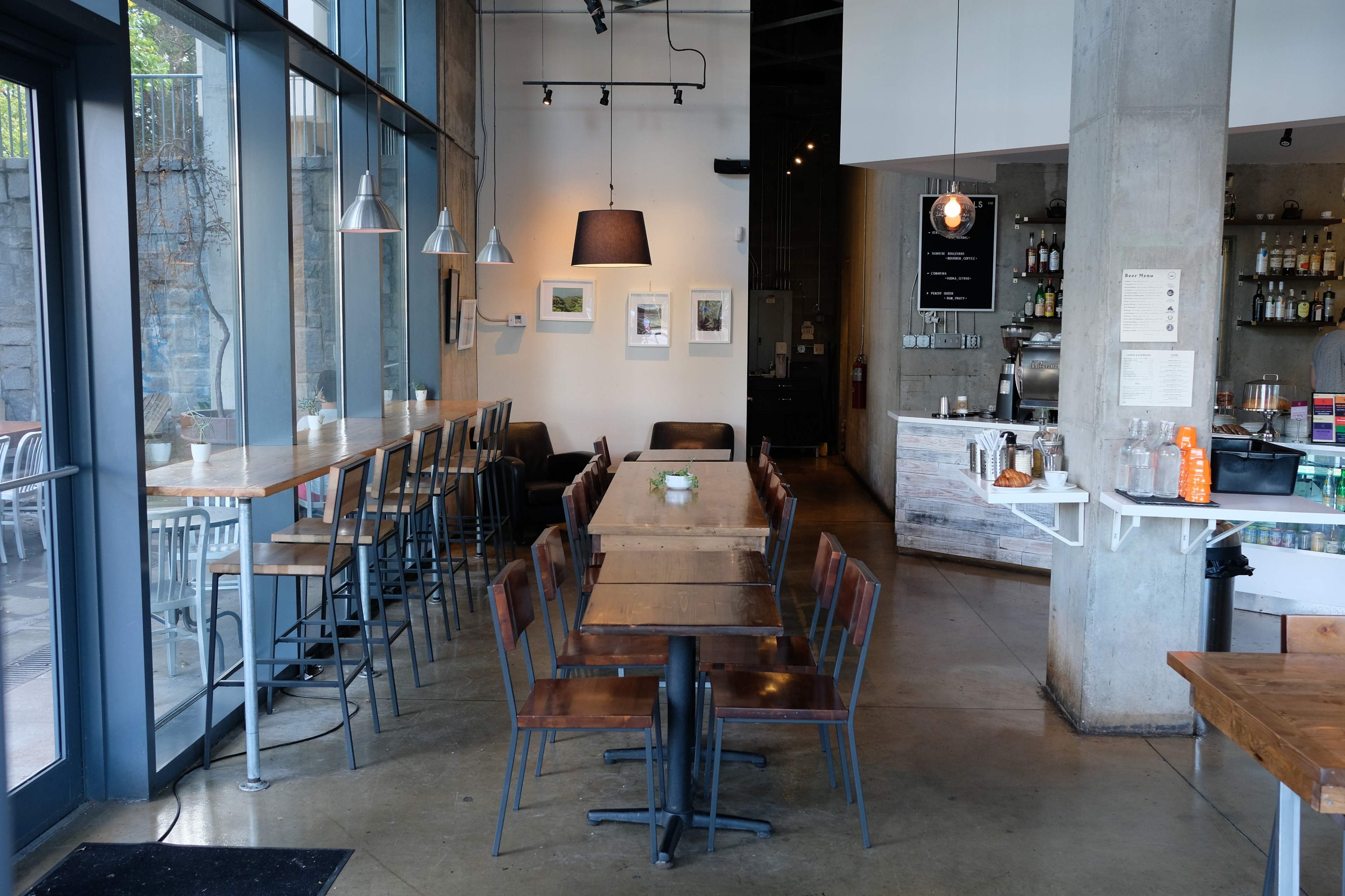 A modern café interior with a mix of high and standard seating arrangements, large windows, and a service counter in the background.