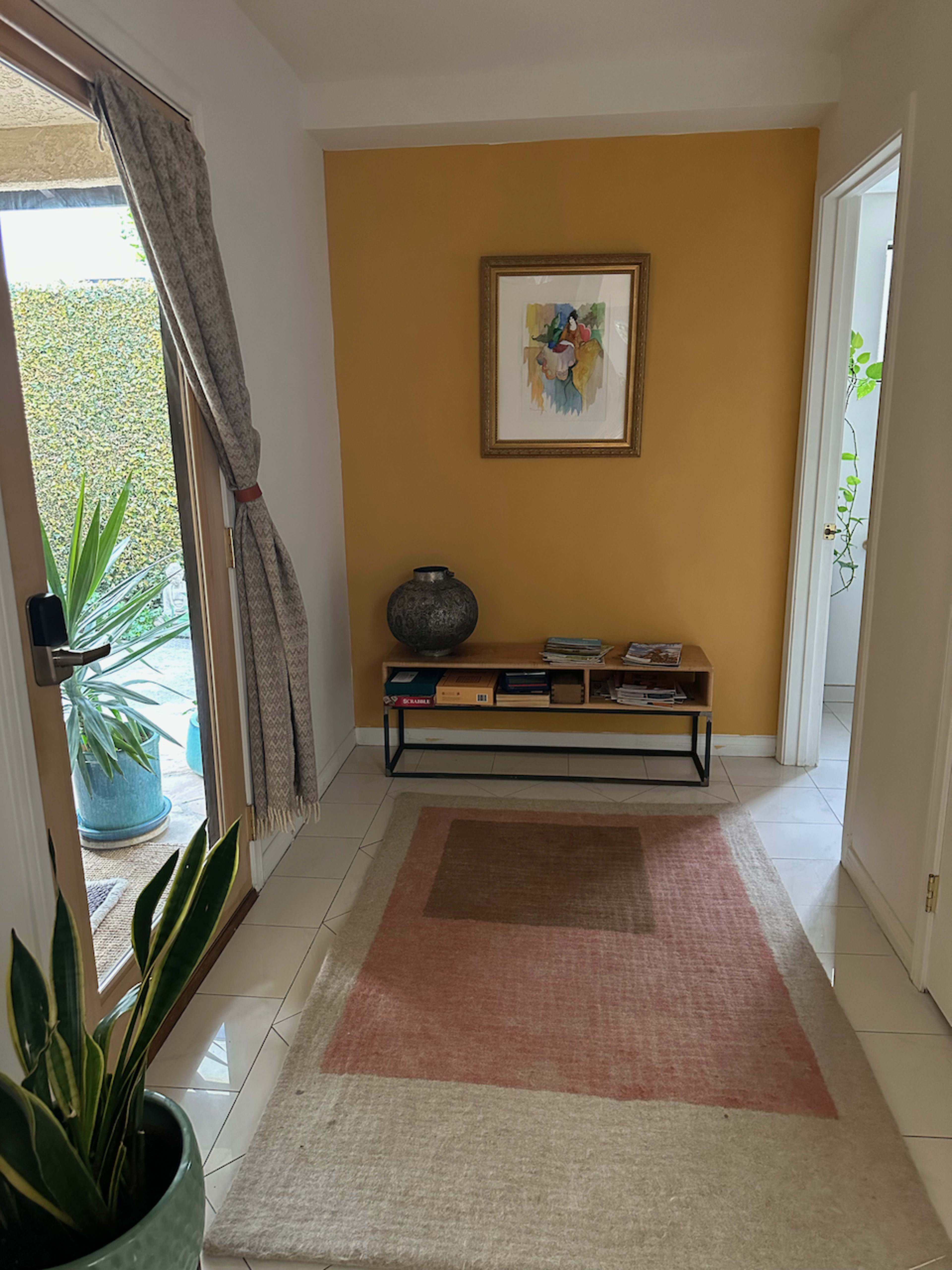 The image shows a hallway with a yellow wall featuring a framed artwork, a wooden shelf with books, and a large potted plant beside a glass door.