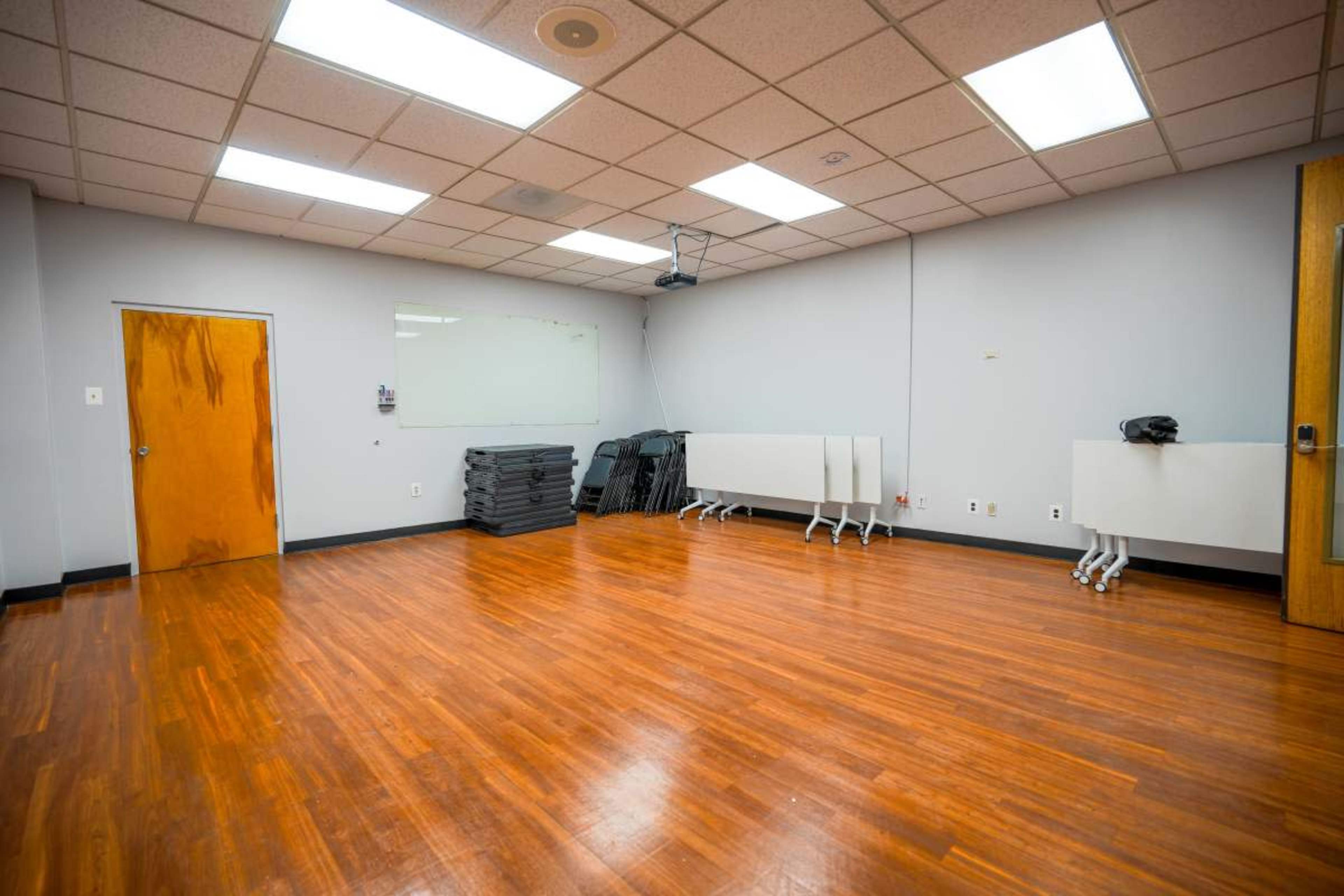 The image shows a sparsely furnished room with a wooden floor, equipped with a wall-mounted projector and several folding tables stacked against the wall.
