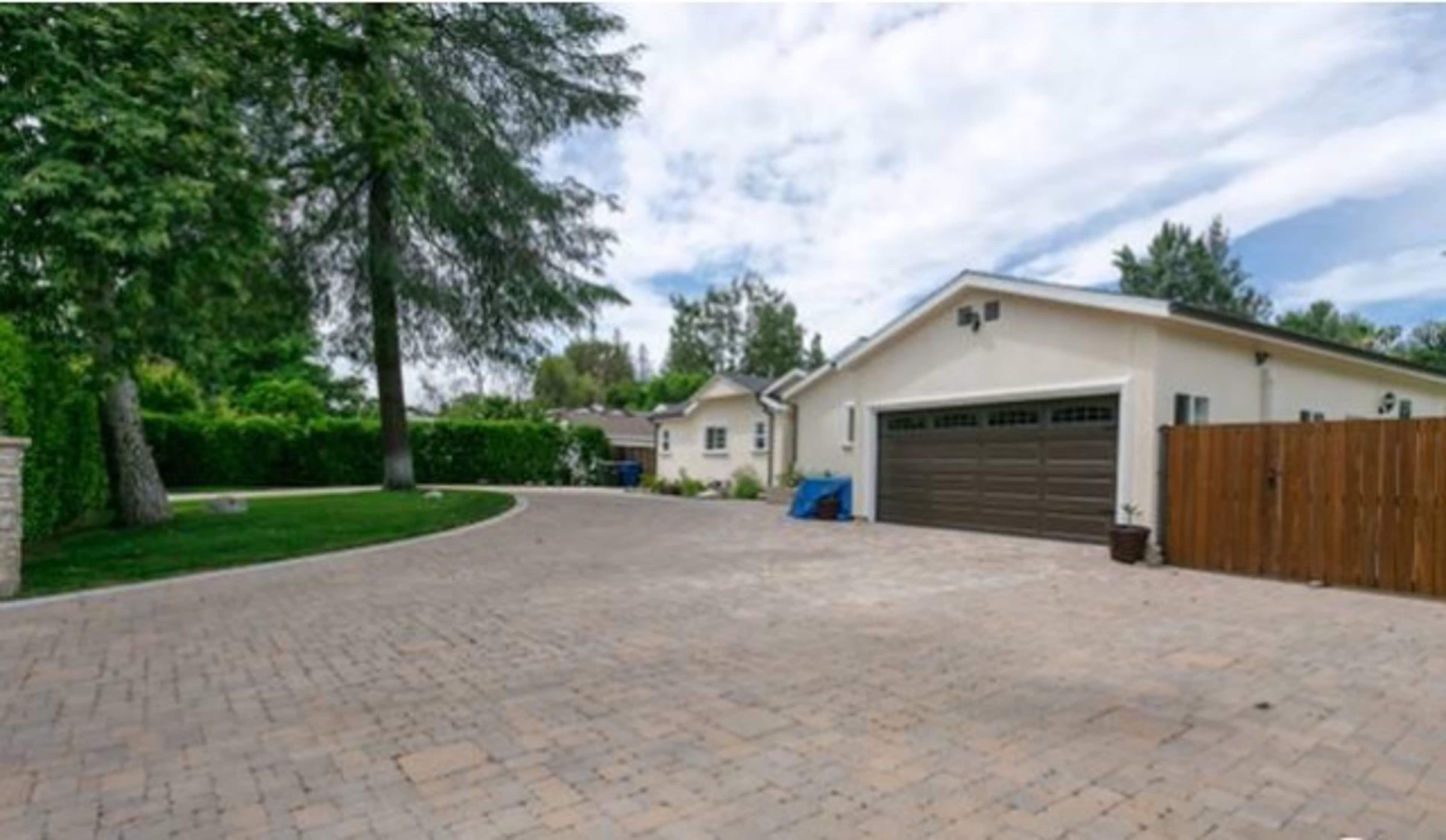 A brick-paved driveway leads to a beige house with a dark garage door, surrounded by green trees and hedges.