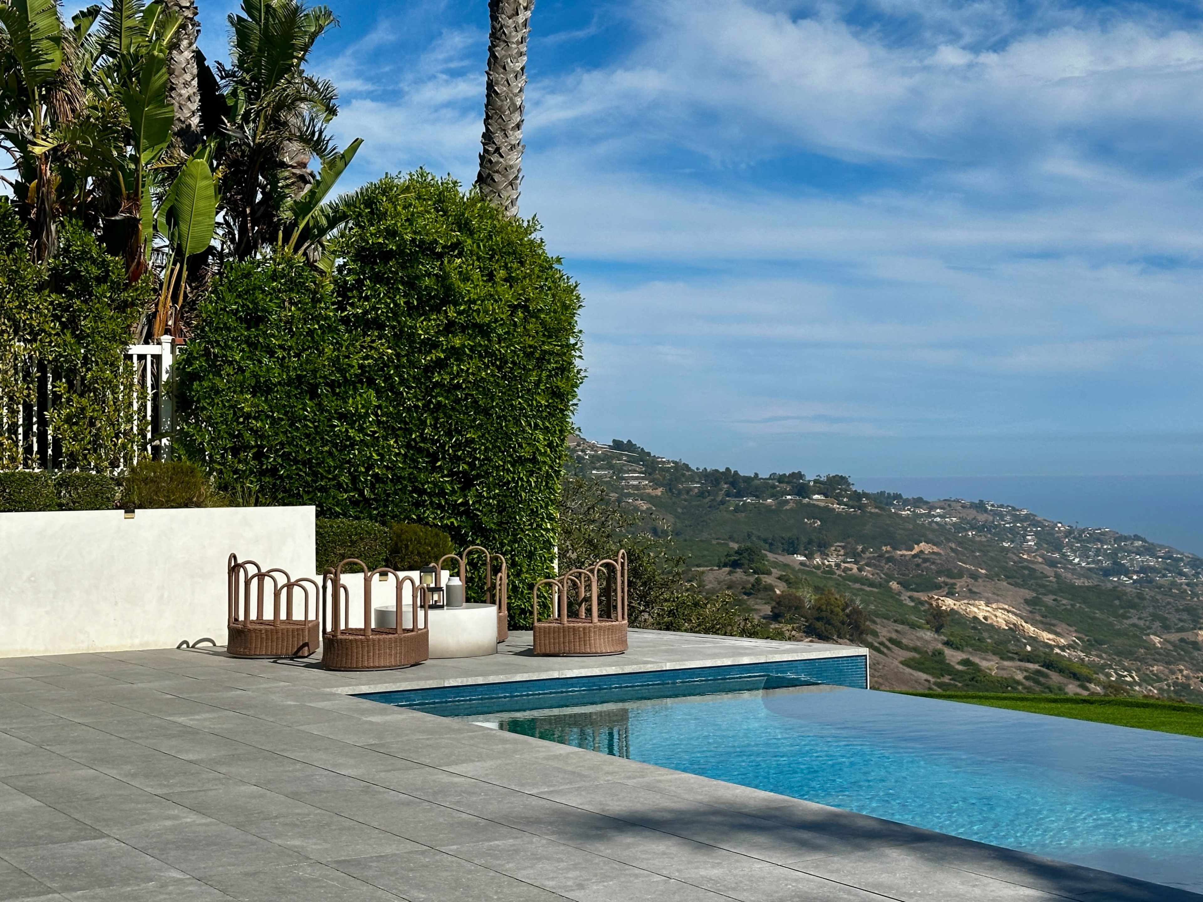 The image shows a luxury outdoor pool area with seating arrangements and landscaped greenery overlooking a hilly coastline under a blue sky.