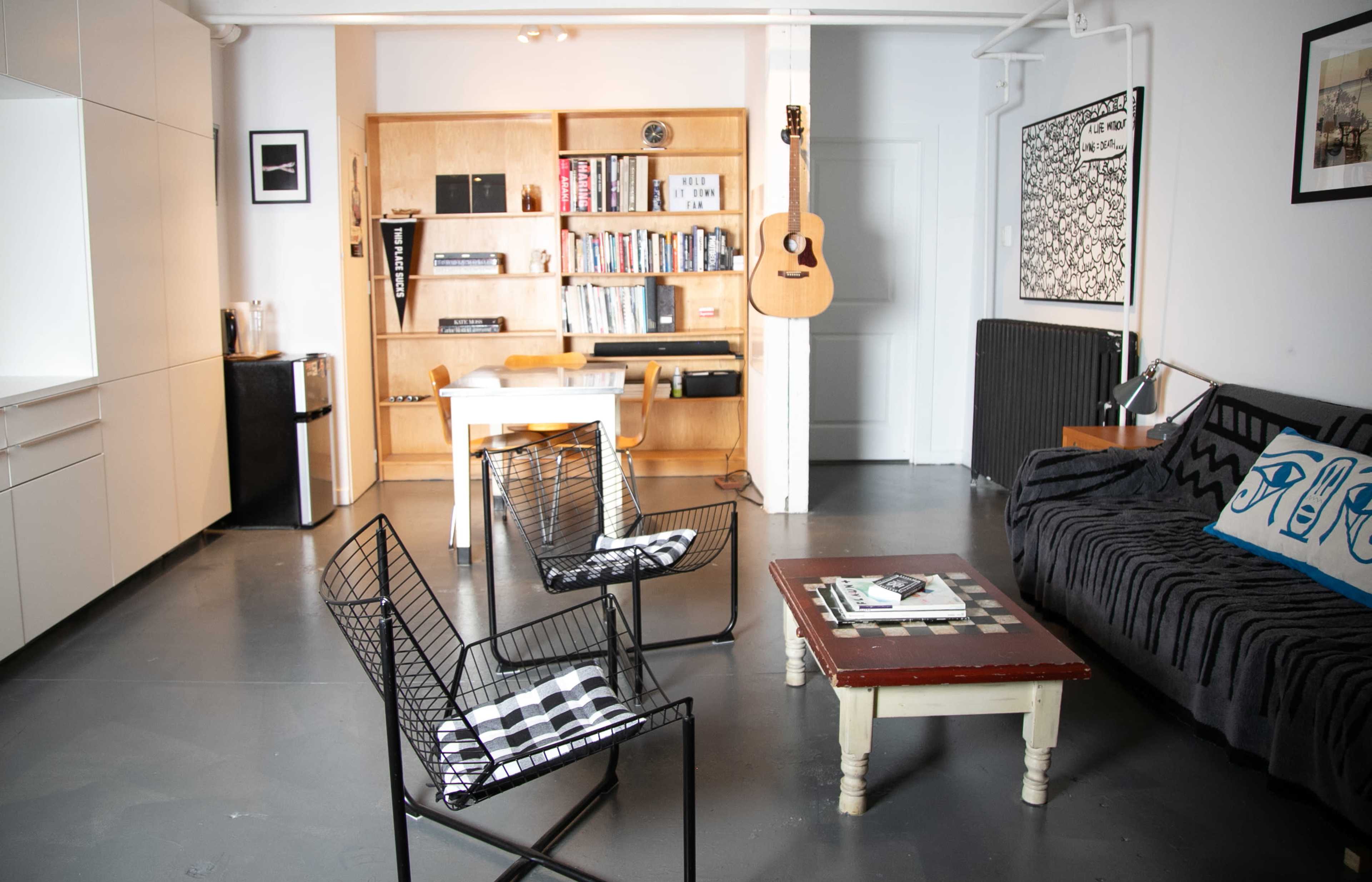 The image shows a modern living space featuring a black couch, a wooden table with chairs, bookshelves, and a guitar hanging on the wall.