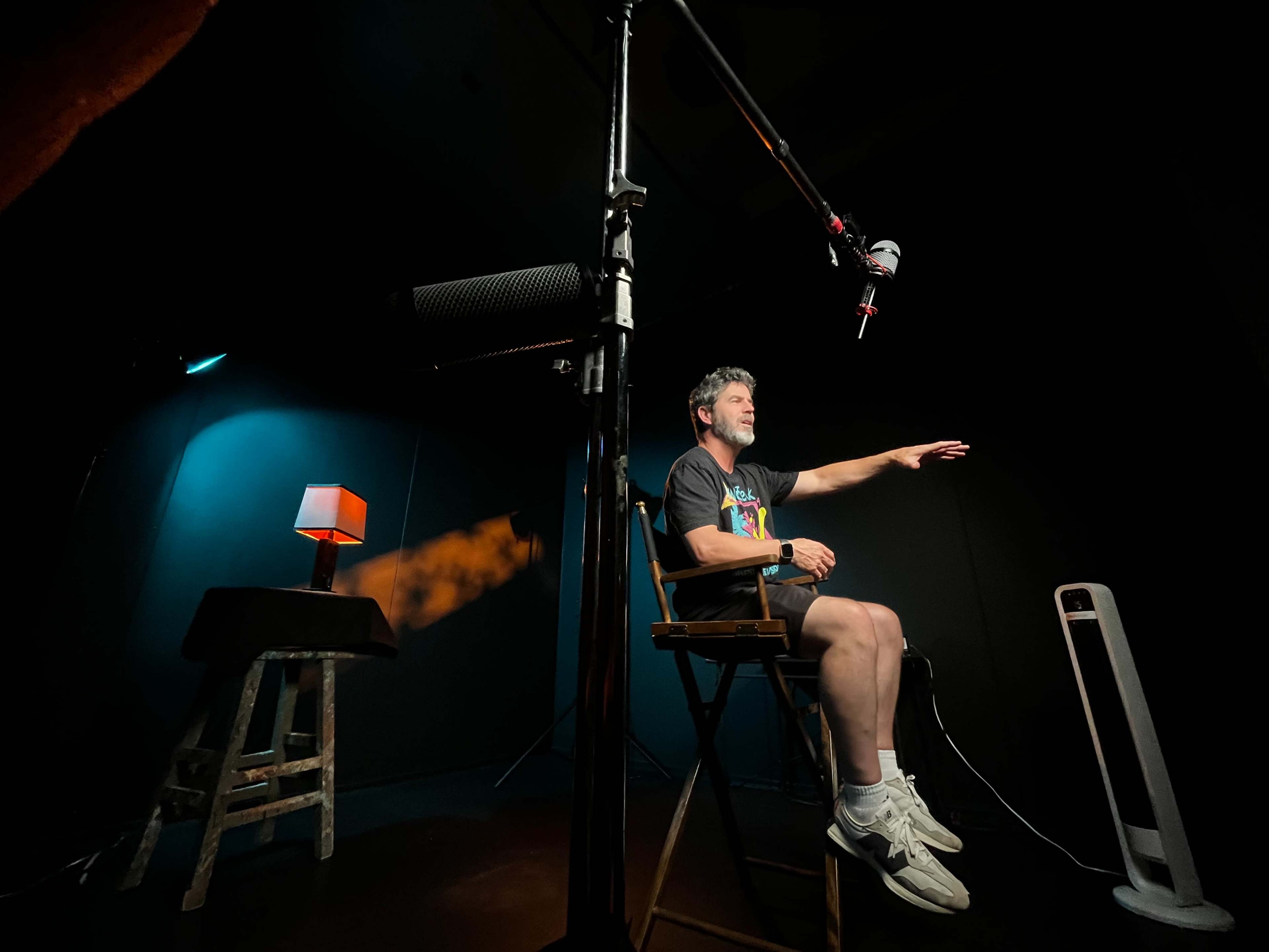 A man sits on a director's chair in a dimly lit studio, gesturing while framed by a microphone and a lamp on a nearby table.