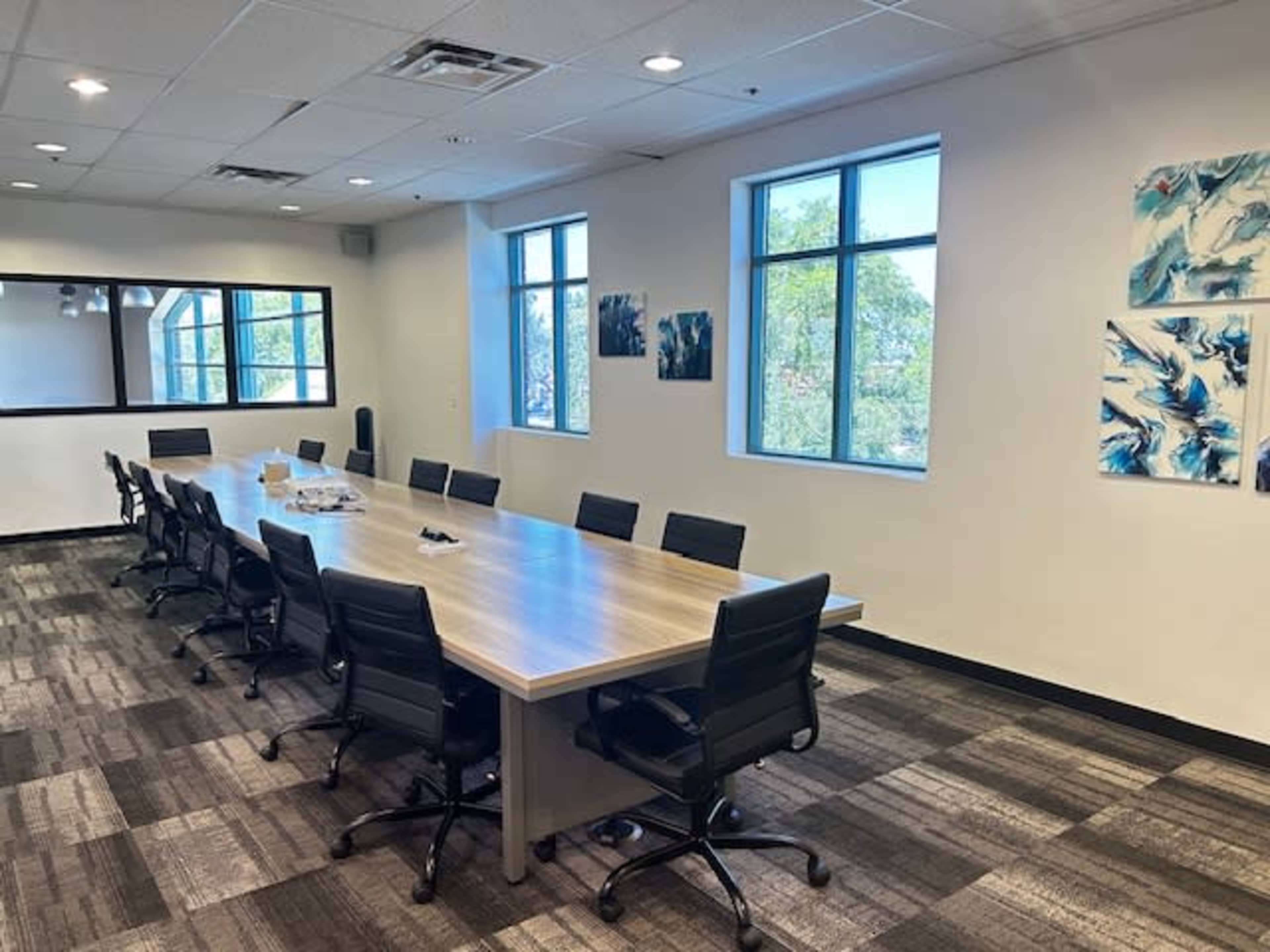 A large conference room features a long wooden table surrounded by black chairs, with windows providing natural light and artwork on the walls.