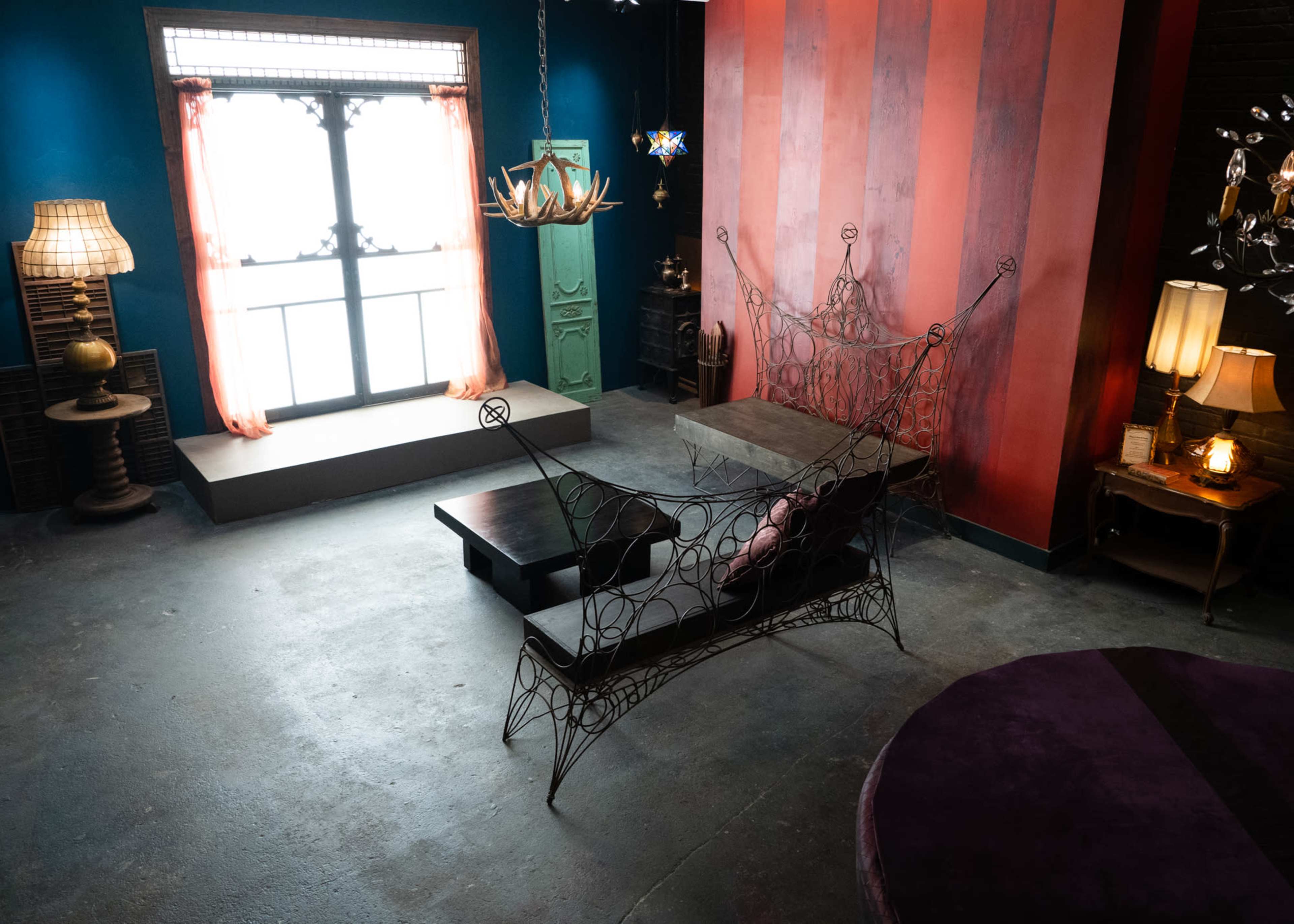 The image shows a stylish room featuring a metal wire sofa, a large round purple rug, and various lighting fixtures against a backdrop of colorful walls and a large window.