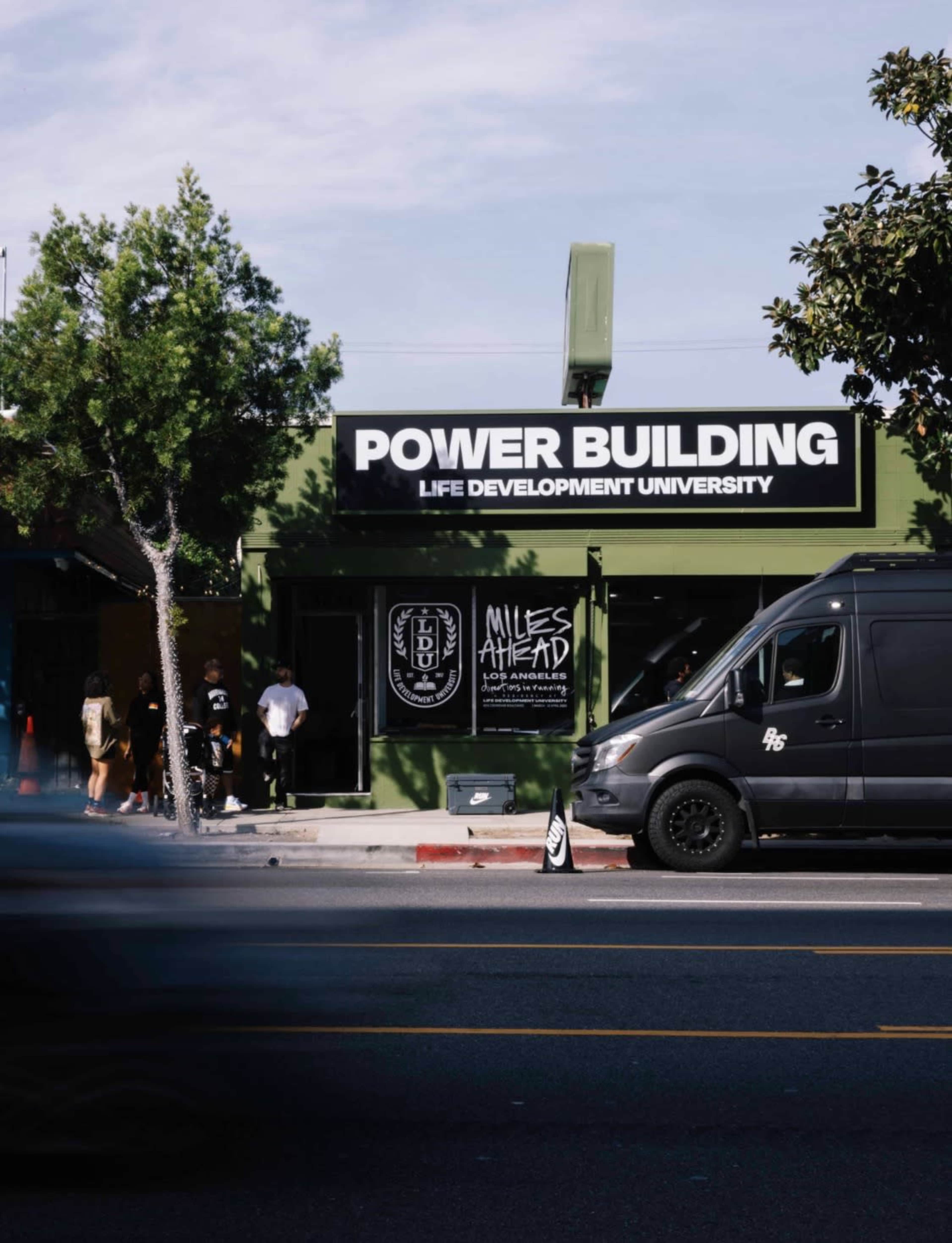 The image shows a large sign for "POWER BUILDING" and "LIFE DEVELOPMENT UNIVERSITY" on a green building, with a van parked nearby and people gathered in front.