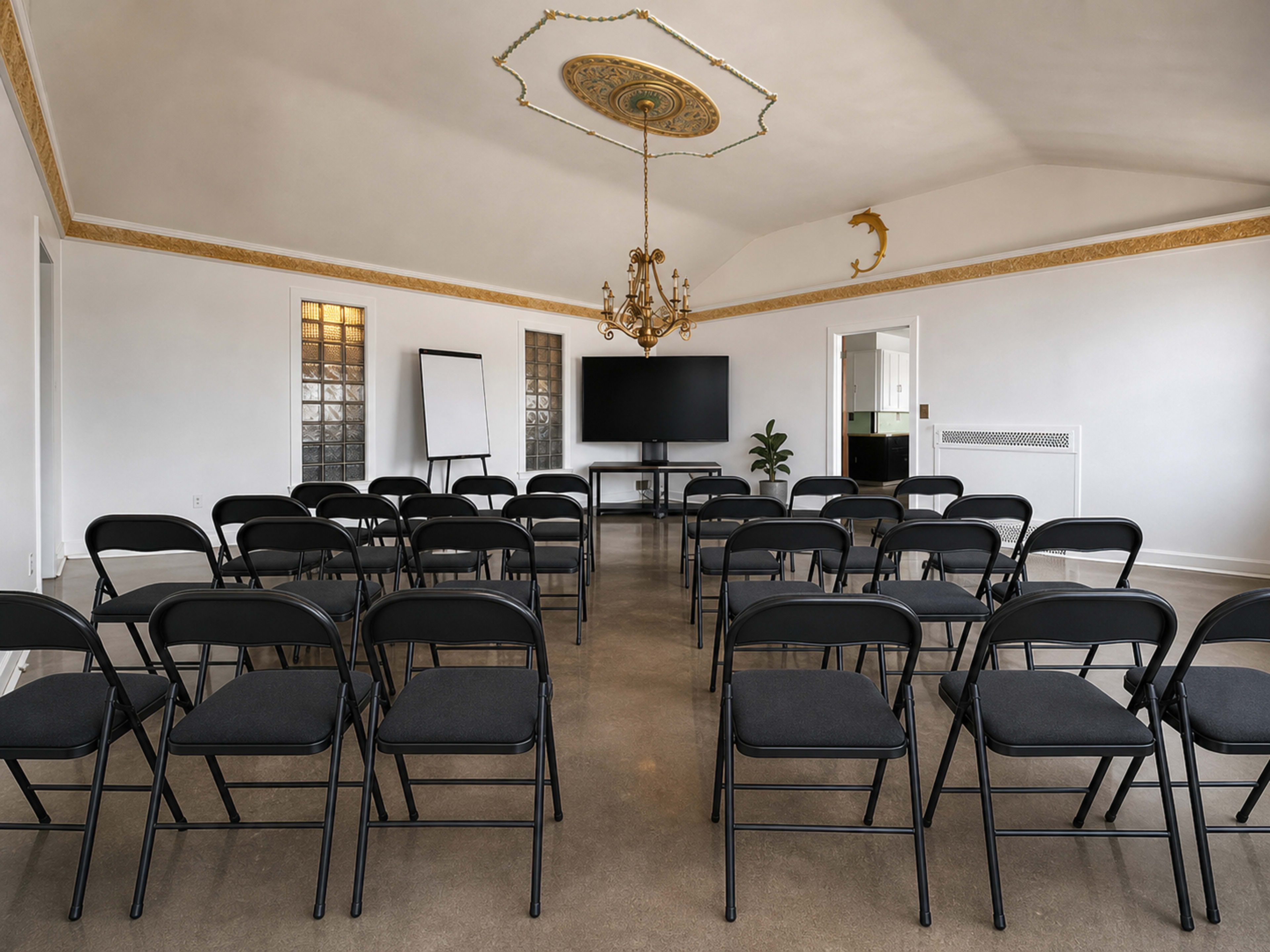 A neatly arranged conference room features rows of black folding chairs facing a large screen and flip chart, under a decorative chandelier.