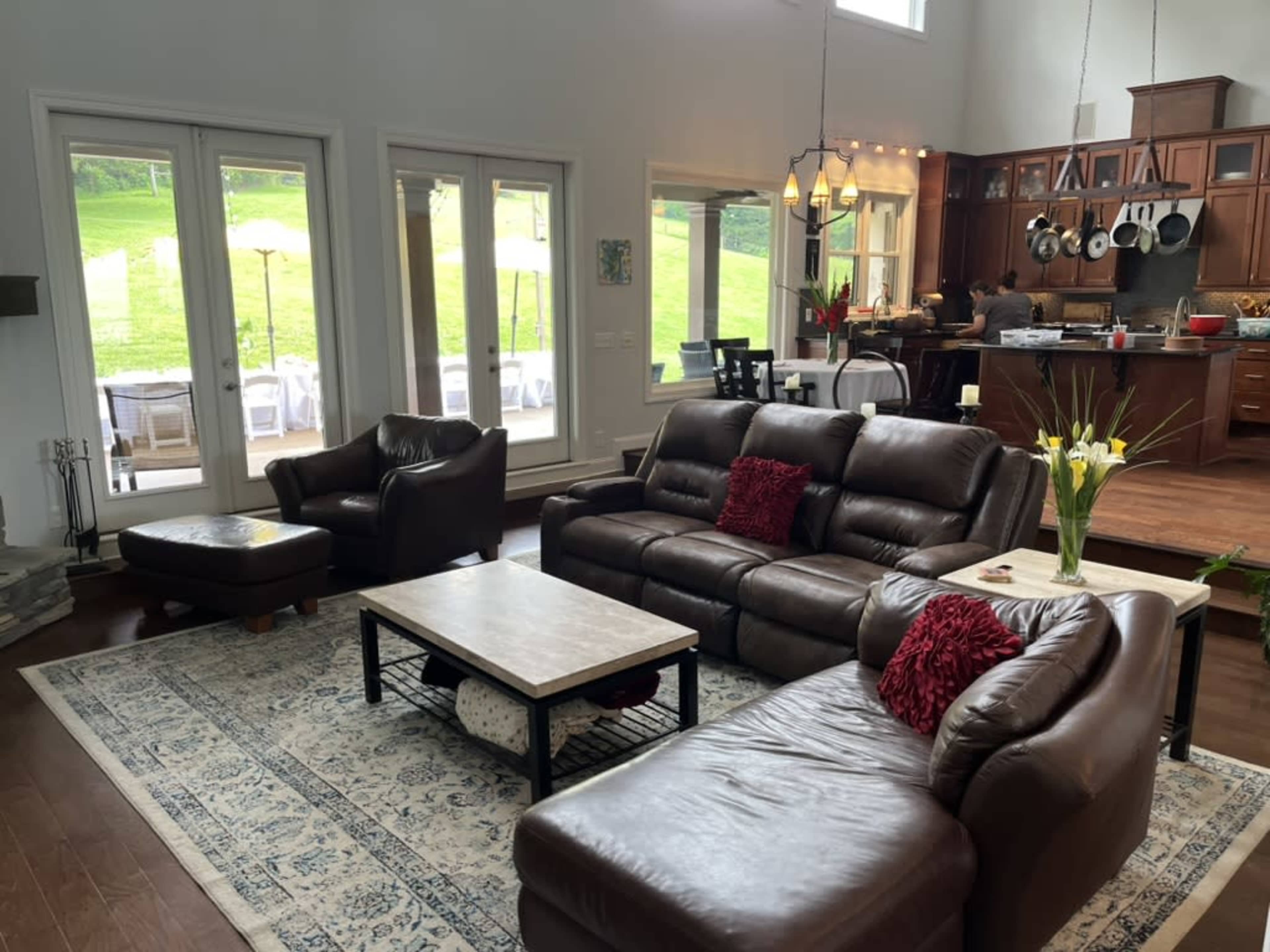 A spacious living room features leather sofas, a coffee table, and large windows overlooking a backyard, with an open kitchen visible in the background.