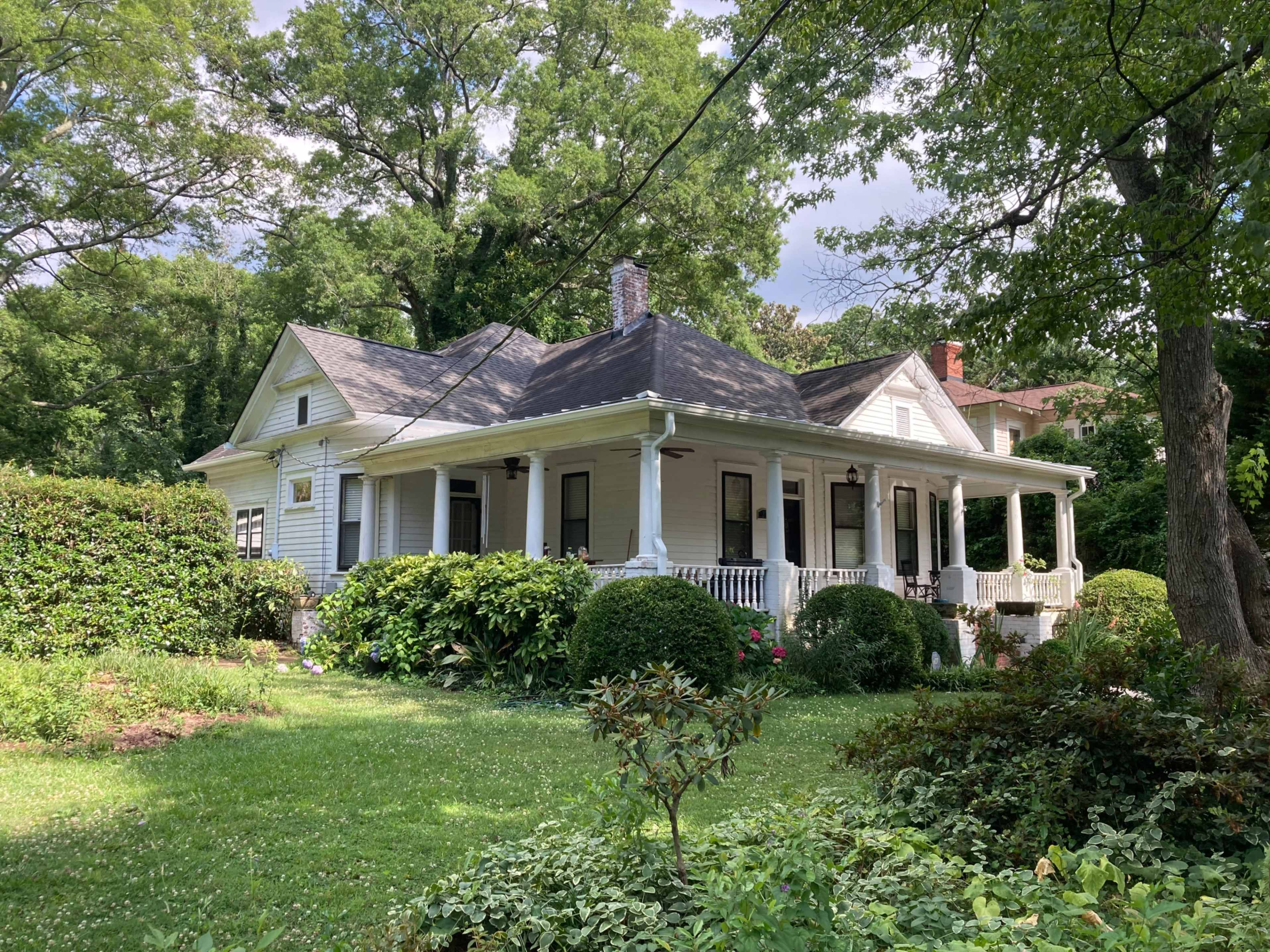 A white, two-story house with a wraparound porch is surrounded by lush greenery and shrubs in a residential area.