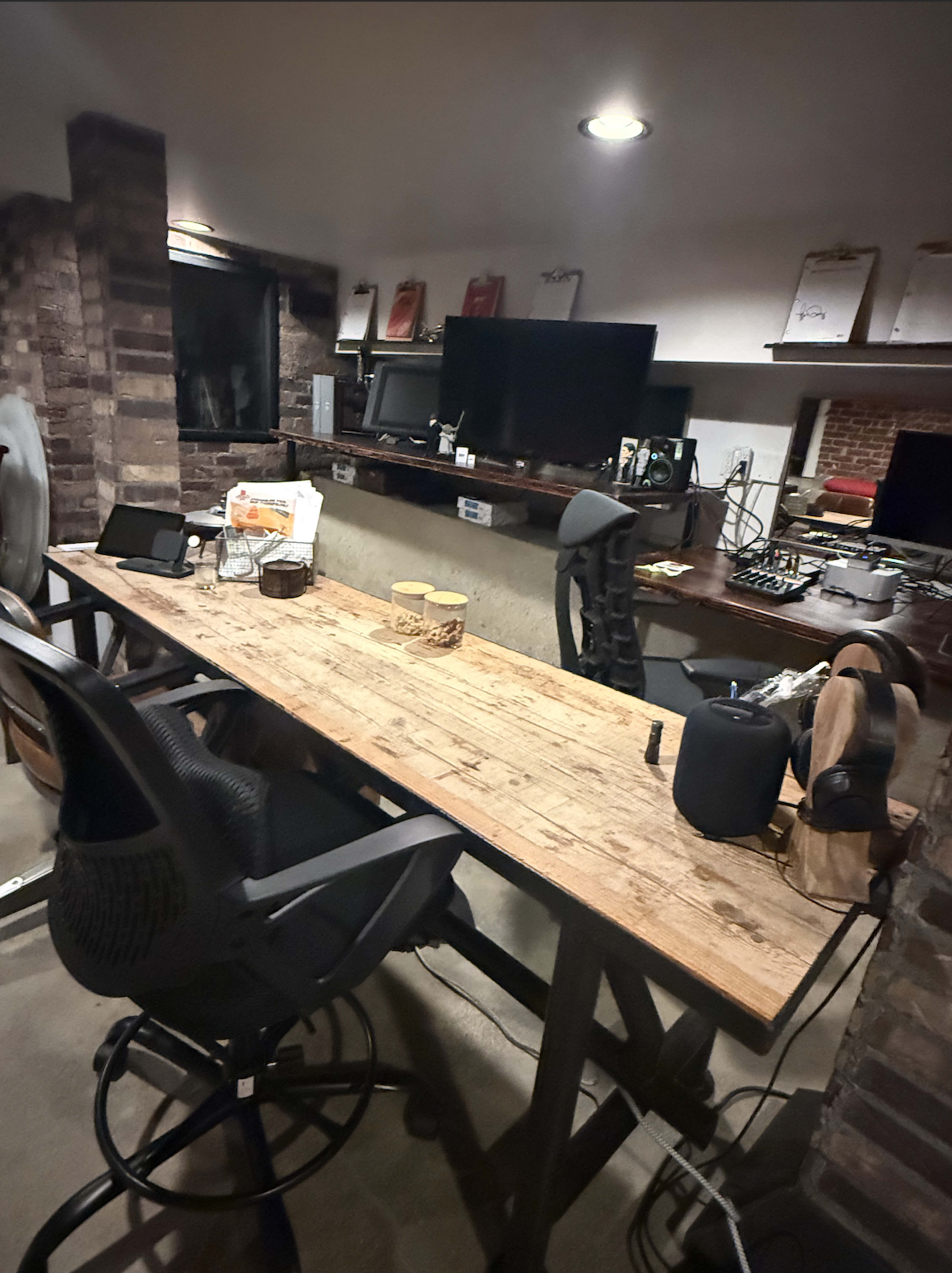 The image shows a modern workspace featuring a large wooden table, two chairs, multiple computer screens, and various items scattered across the surface.
