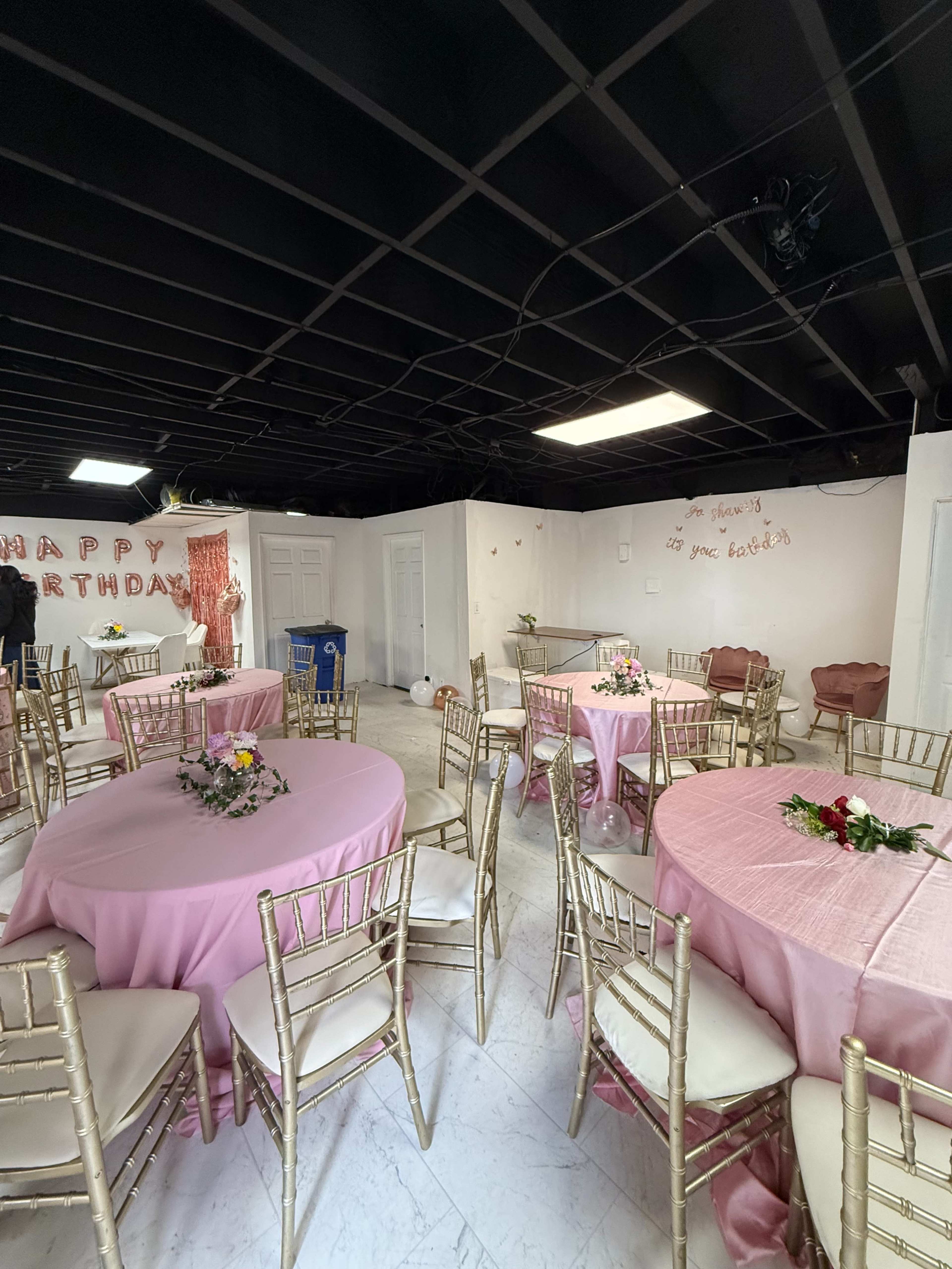 The image shows a decorated indoor event space with round tables covered in pink tablecloths and gold chairs, featuring floral centerpieces and a "Happy Birthday" banner on the wall.