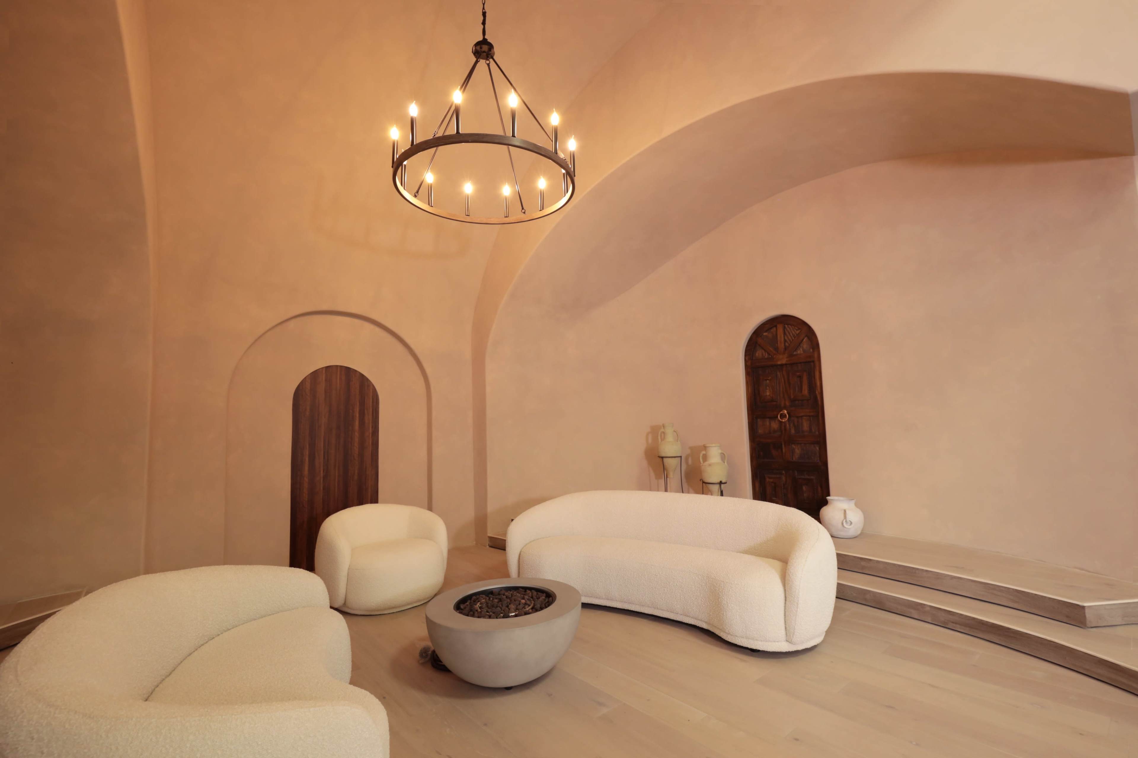 A modern, minimalist lounge area with two curved sofas around a circular coffee table, all set beneath a chandelier in an arched room.