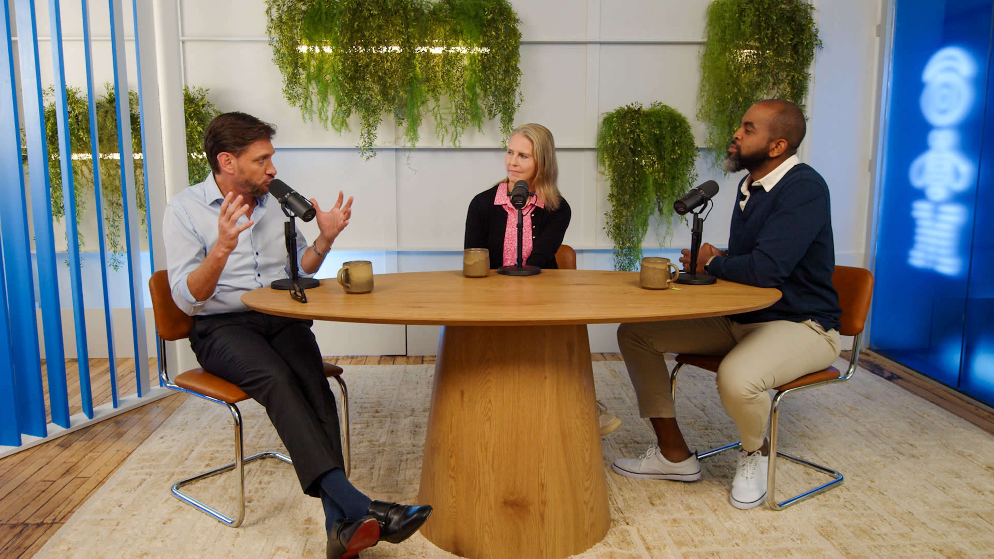 Three people are seated around a round wooden table in a podcast studio, engaged in discussion with microphones in front of them.
