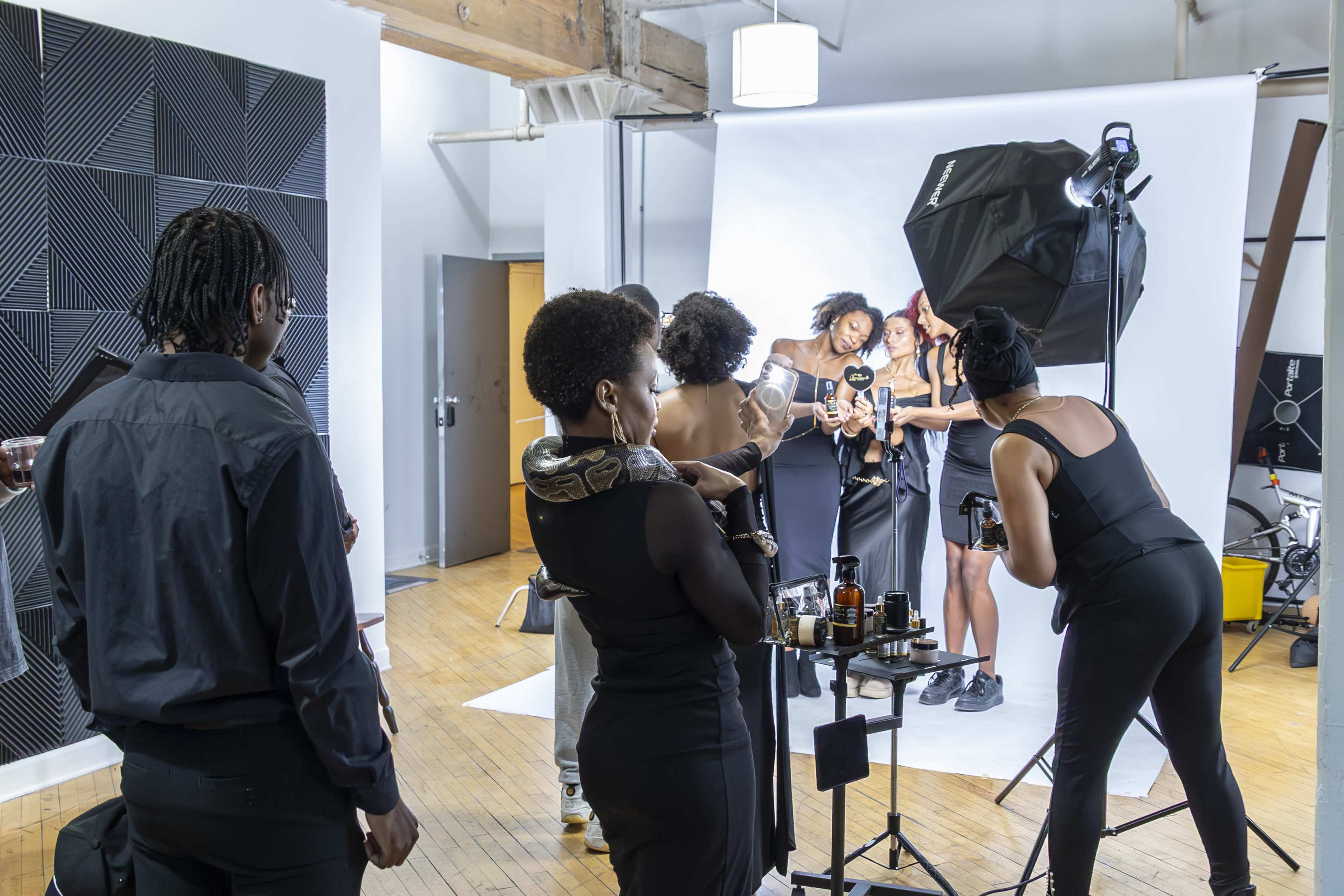 A group of five women poses for a photo shoot in a studio, while several individuals assist with lighting and equipment.