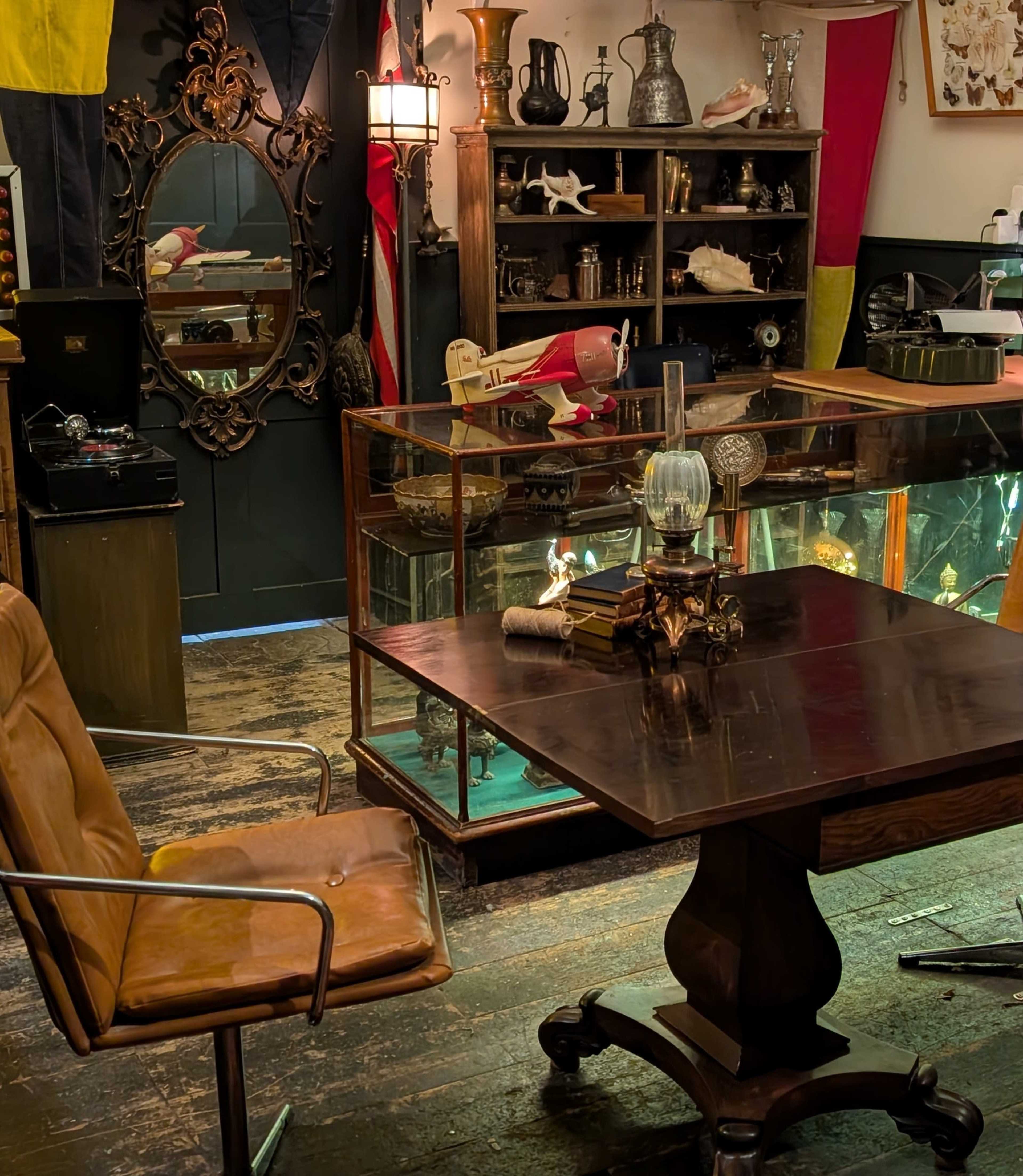 The image shows an antique shop interior featuring a wooden table, a leather chair, and various historical artifacts displayed in glass cases against a backdrop of flags and shelves filled with collectibles.