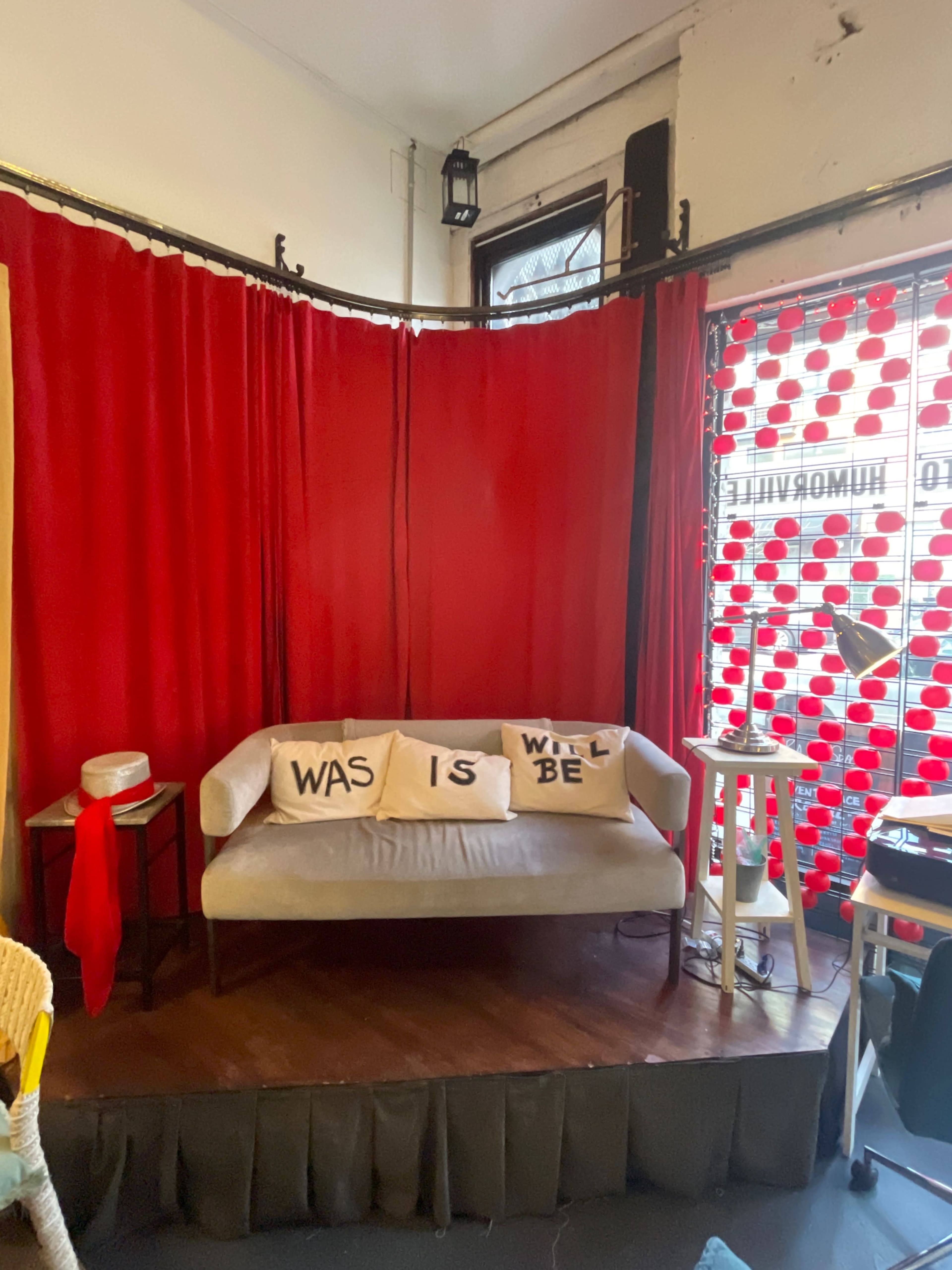 The image shows a cozy seating area featuring a light-colored sofa adorned with decorative pillows that read "WAS," "IS," and "WILL BE," against a backdrop of red curtains and a patterned window.
